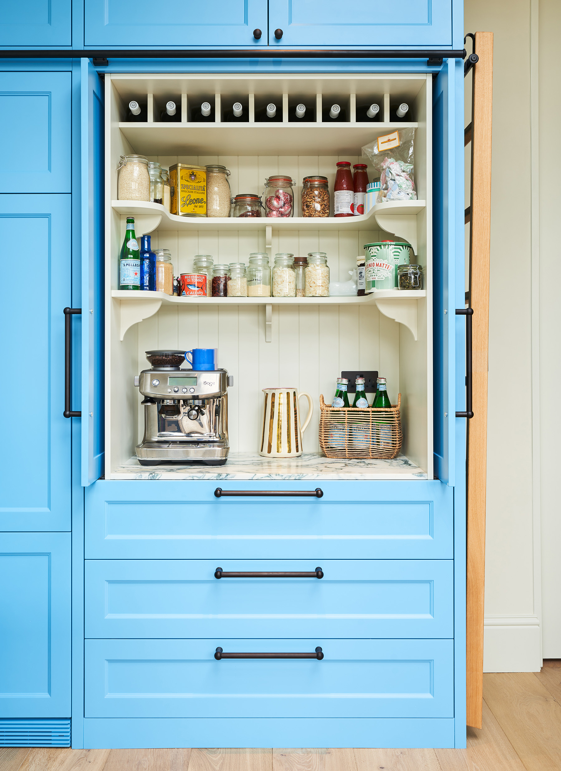 Bright blue kitchen coffee bar cabinet with open bi-fold doors, white tongue-and-groove larder interior, stainless steel espresso machine, shelves with glass jars, and wine cubbies above drawers
