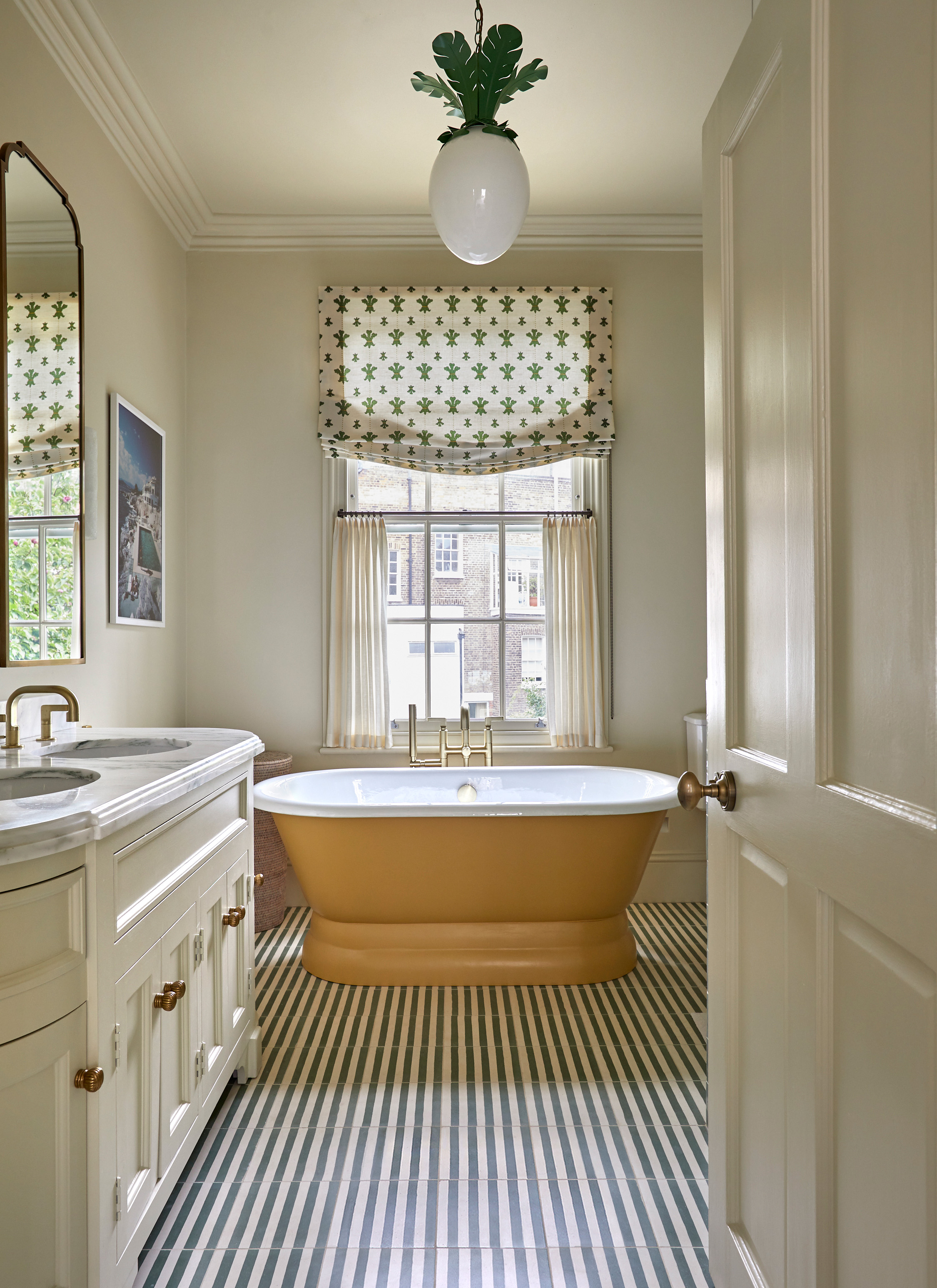 Cream bathroom with a yellow freestanding soaking tub set against a window with patterned blinds. A marble-topped double vanity sits to the left, paired with striped floor tiles and a whimsical pendant light overhead