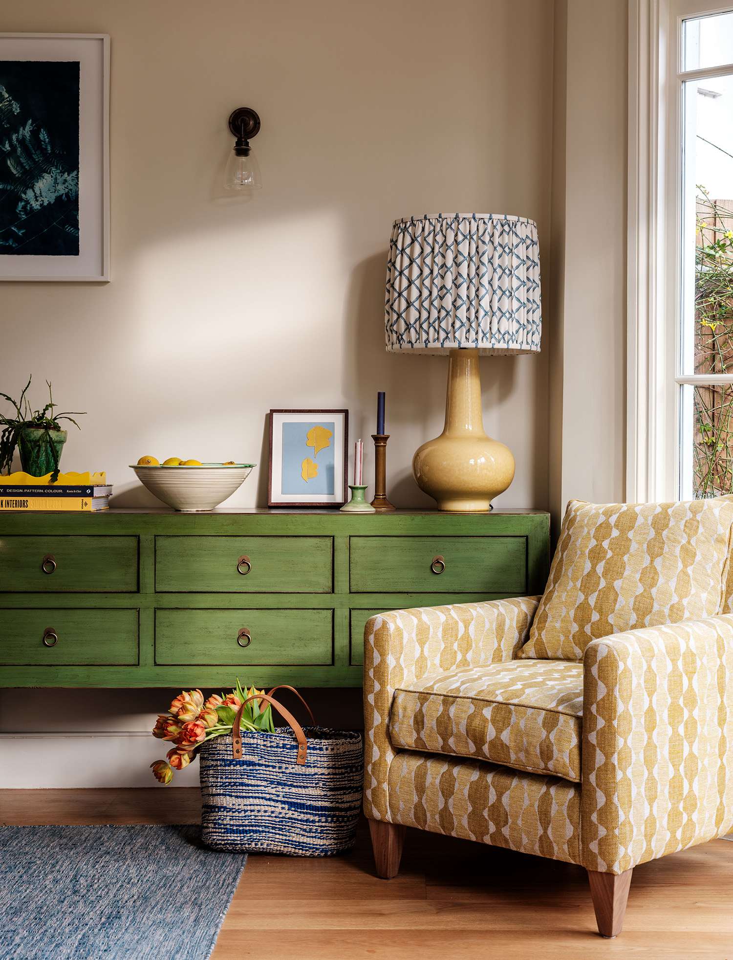 A close up of a jade green sideboard with six drawers, topped with a yellow table lamp with a fabric shade, candlesticks, picture frame and large bowl of lemons, in front is a patterned armchair in shades of yellow, and a blue speckled jute bag with tulips