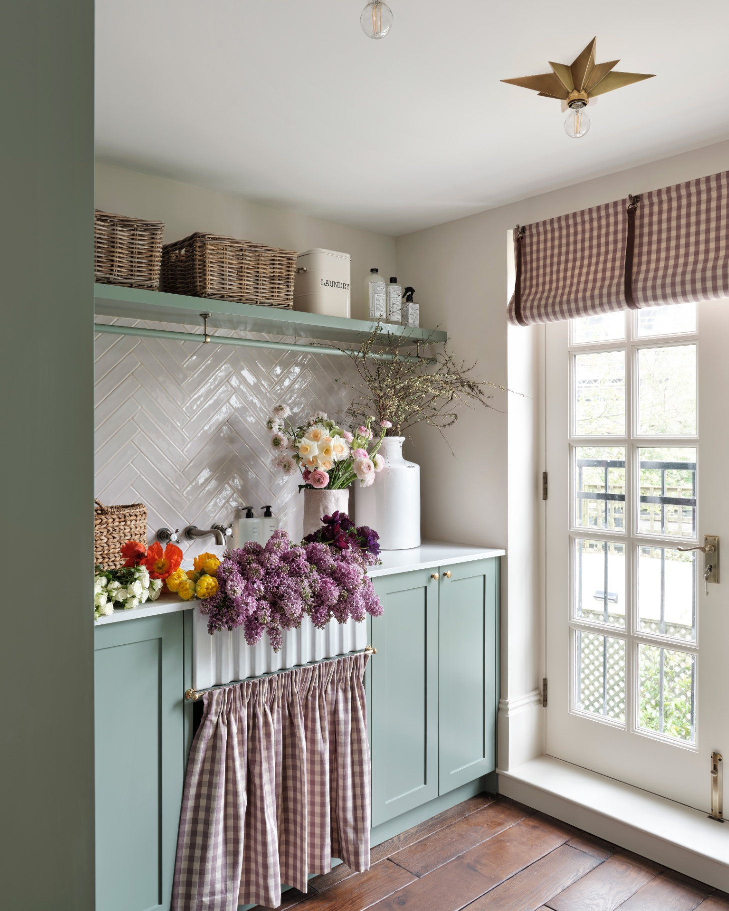 a utility room with sage green cabinetry, a lilac and white gingham fabric skirt under a sink filled with floewrs and a high shelf with wicker baskets
