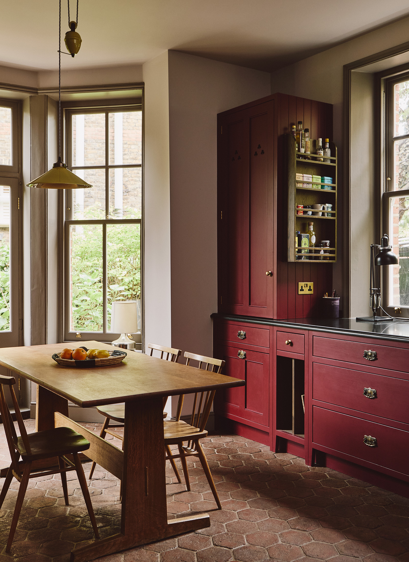 A kitchen with tall ceilings and hexagon stone flooring. To the right is red cabinetry with brass handles and to the left is a wood dining table, topped with a bowl of fruit, and spindle-back chairs. Above hangs a simple ceramic rise and fall pendant. 