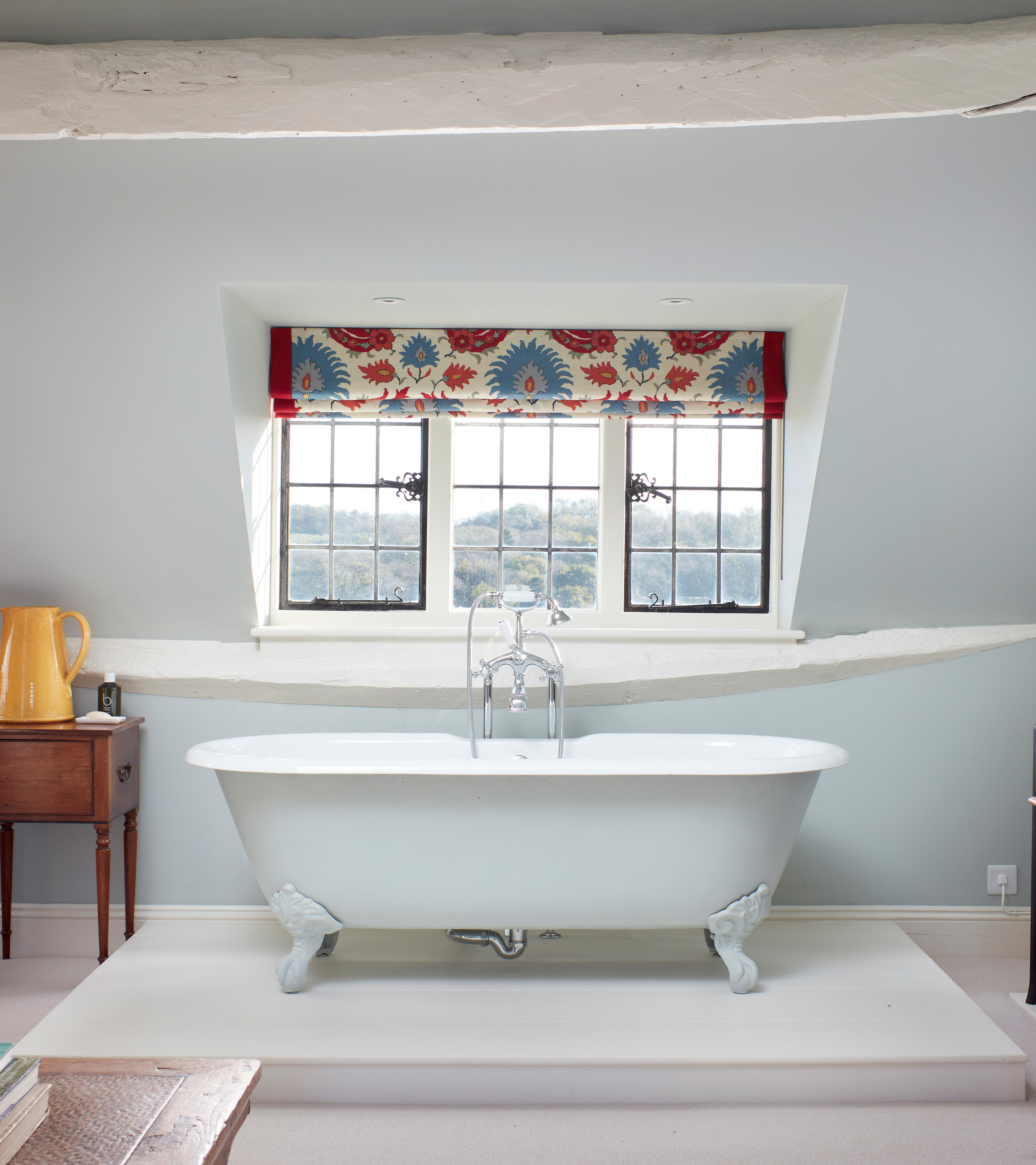 a bathroom with pale blue walls and white beams, a window with a blue and red ikat pattern blind and a freestanding white bath