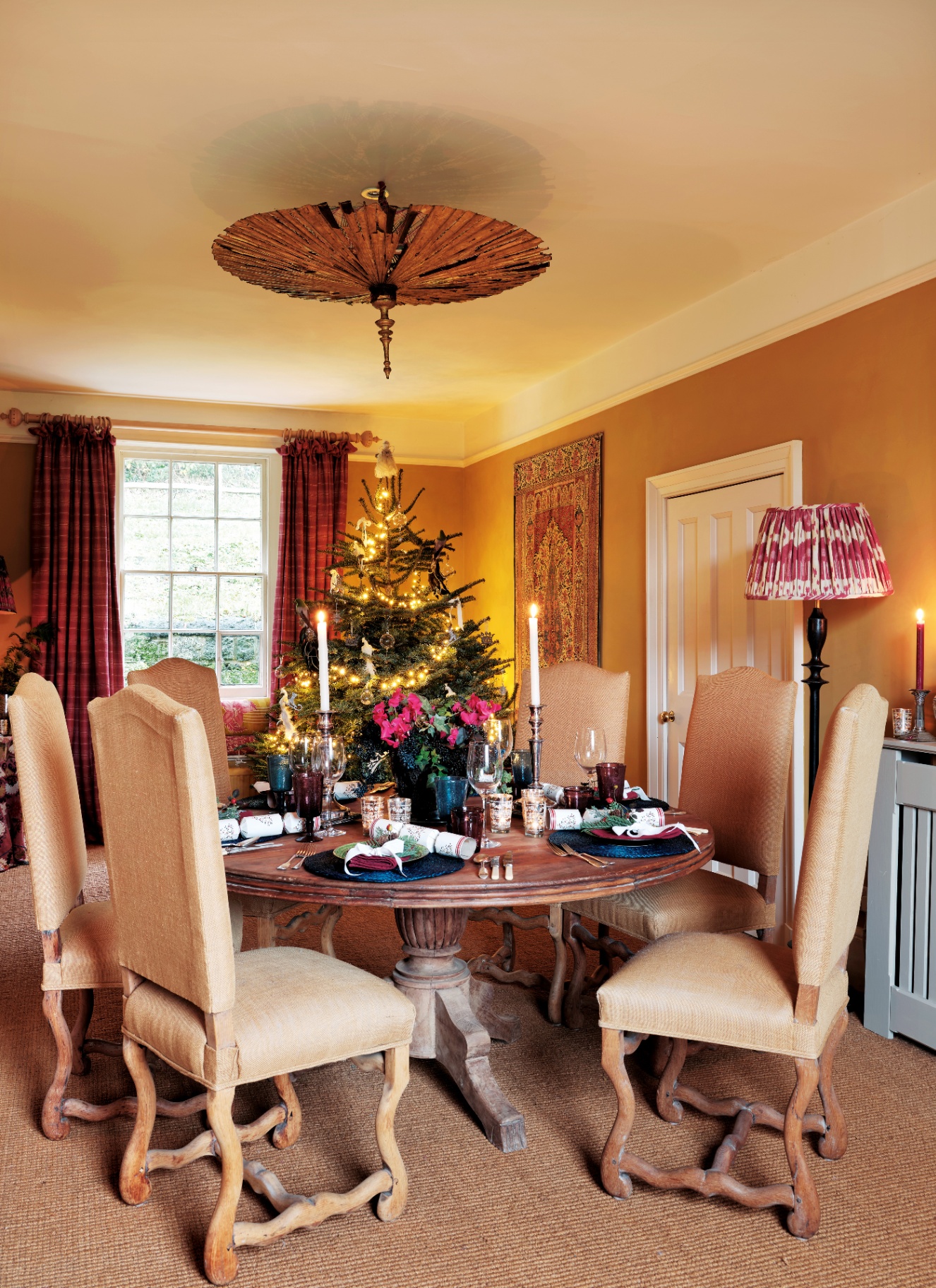 A round wooden dining table laid with tall candles and festive crackers in front of a window with tartan curtains