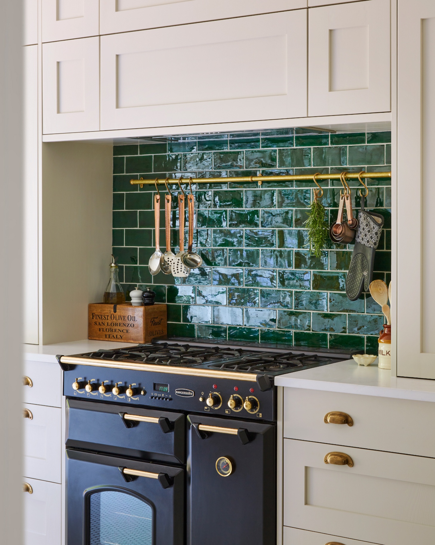 green splashback behind a black range cooker and a gold hanging rail with herbs and copper tools hanging from it
