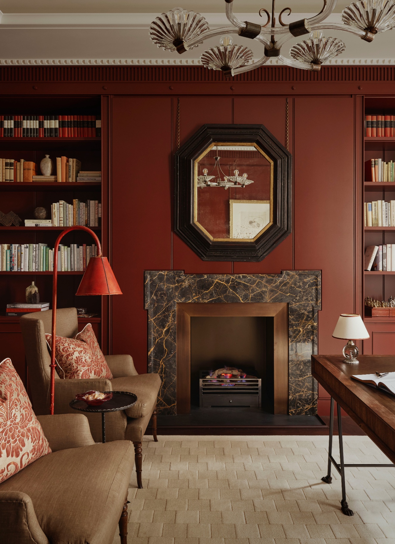a living room with warm chestnut brown walls, a large brown marble fireplace with built in bookcases either side, a glass shell shaped chandelier and two brown armchairs