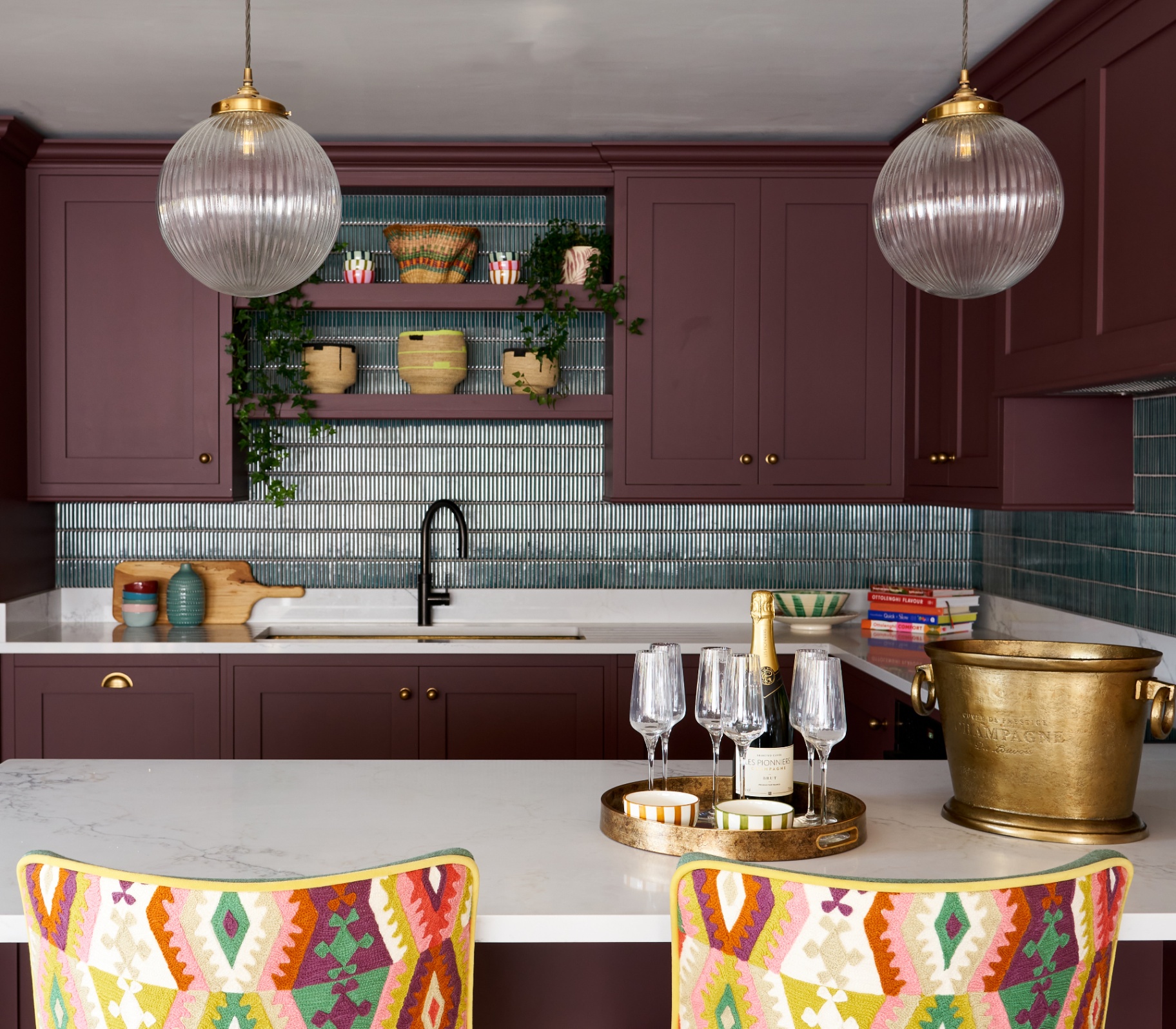 a kitchen with plum purple cabinets, dark teal tiles, two globe glass pendants and two bright coloured ikat print chairs