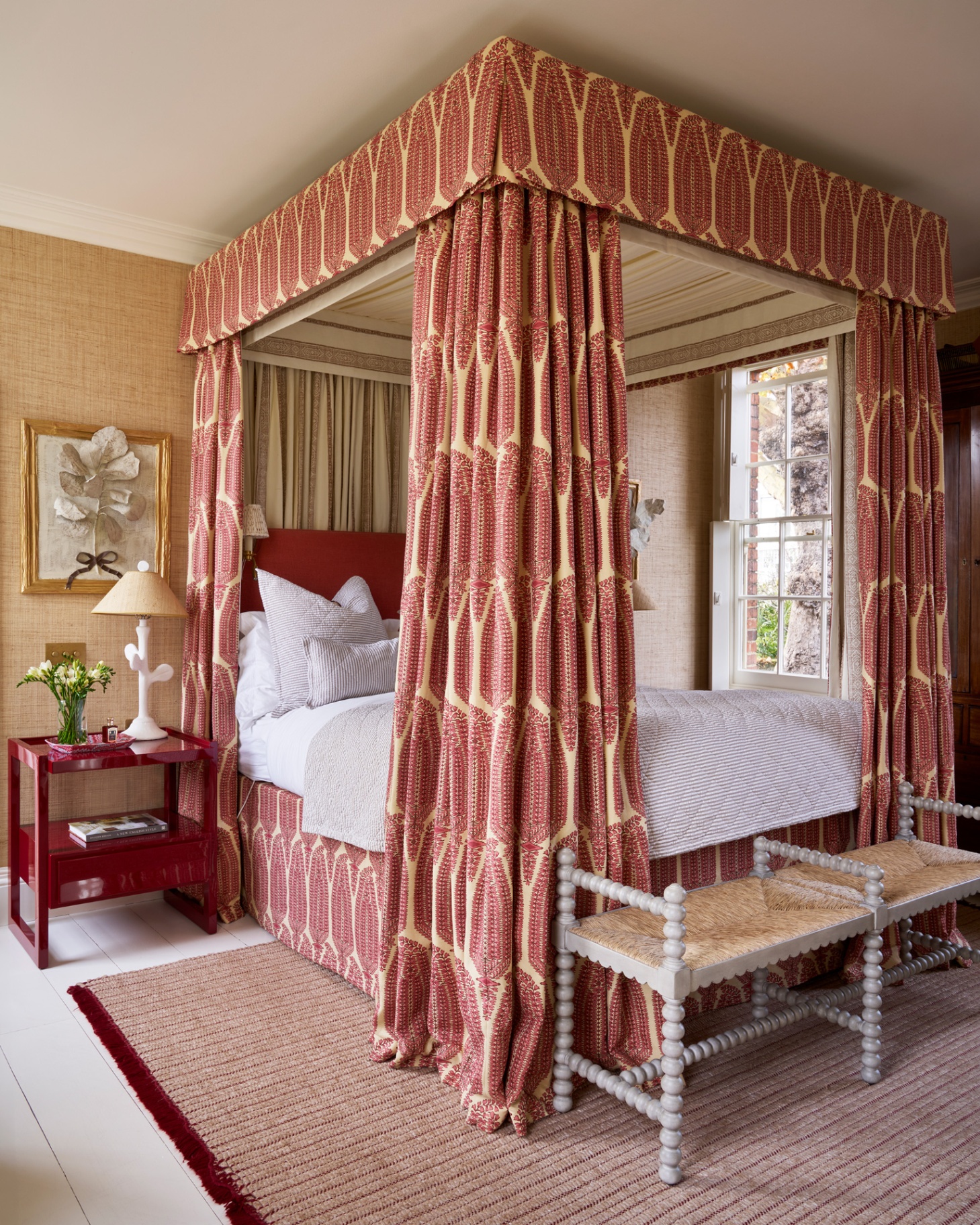 a large four posted bed with red and white patterned drapes, a shiny red bedside table and a grey bobbin bench at the end of the bed