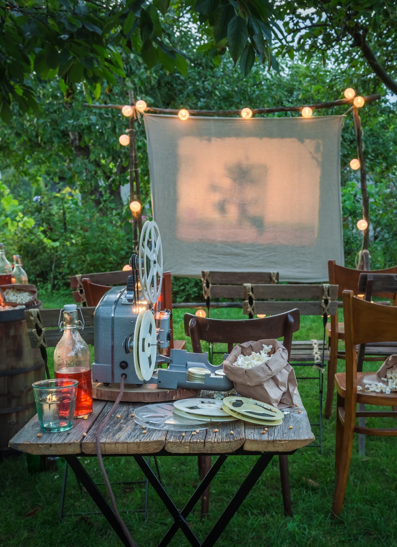 AN OUTDOOR CINEMA WITH CANVAS SCREEN BETWEEN WOODEN POLES WITH FESTOON LIGHTS, WOODEN CHAIRS FACING THE SCREEN AND A WOODEN TABLE WITH A VINTAGE PROJECTOR