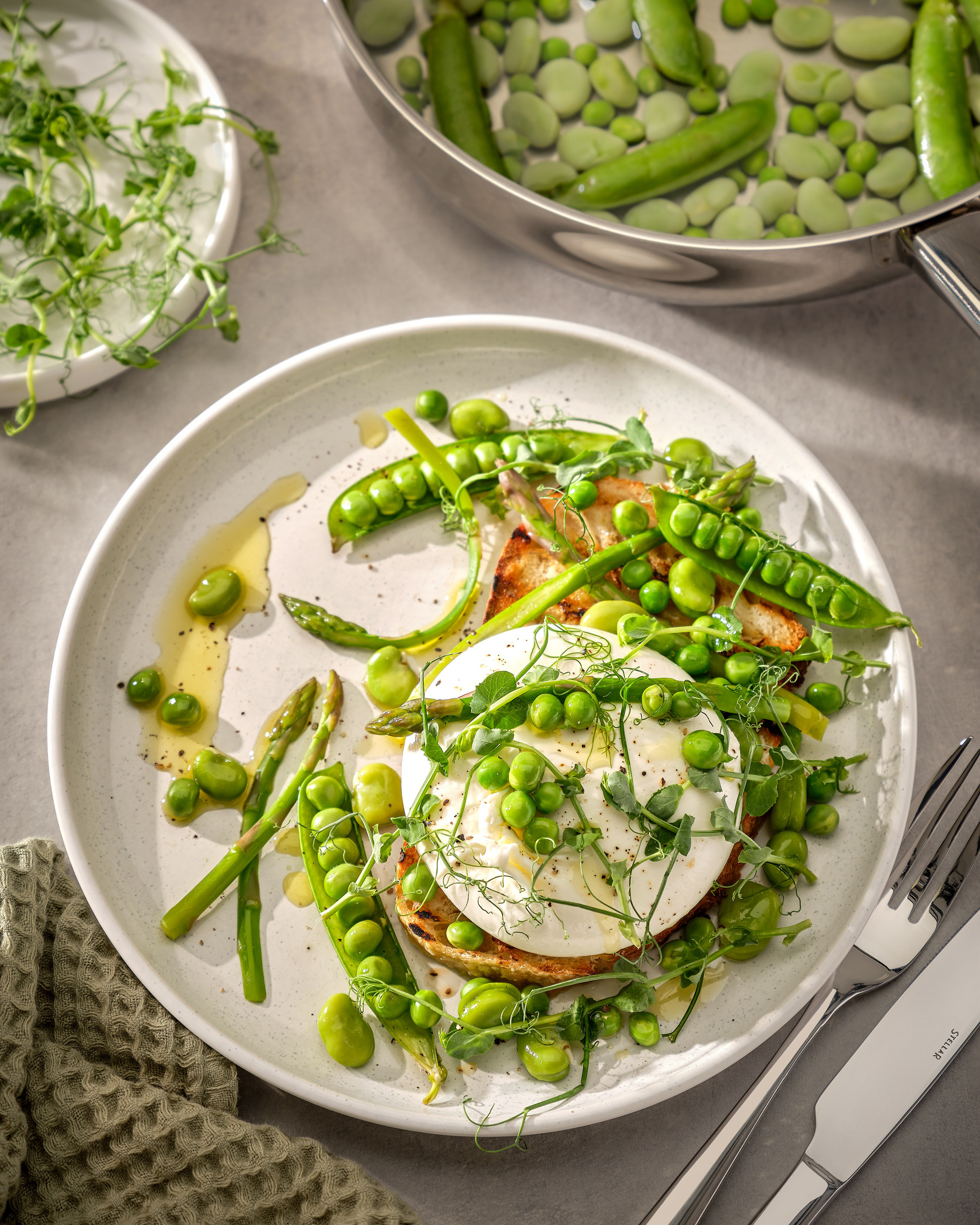 a white plate with sourdough, burratta, broad beans and peas with silver cutlery