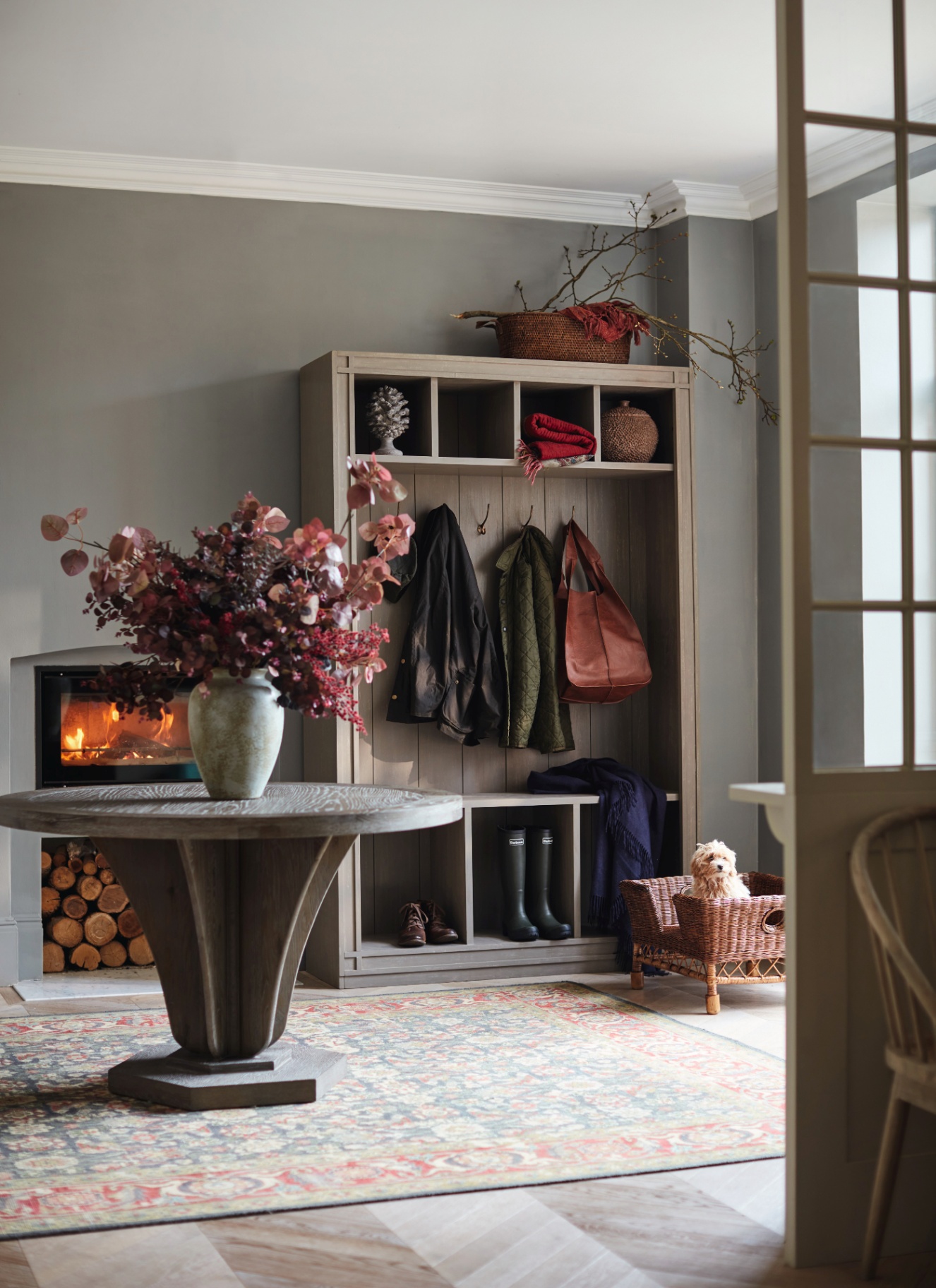 a hallway with a large round table with a vase of red faux leaves, a staorage unit with boots and coats and a rattan dog bed