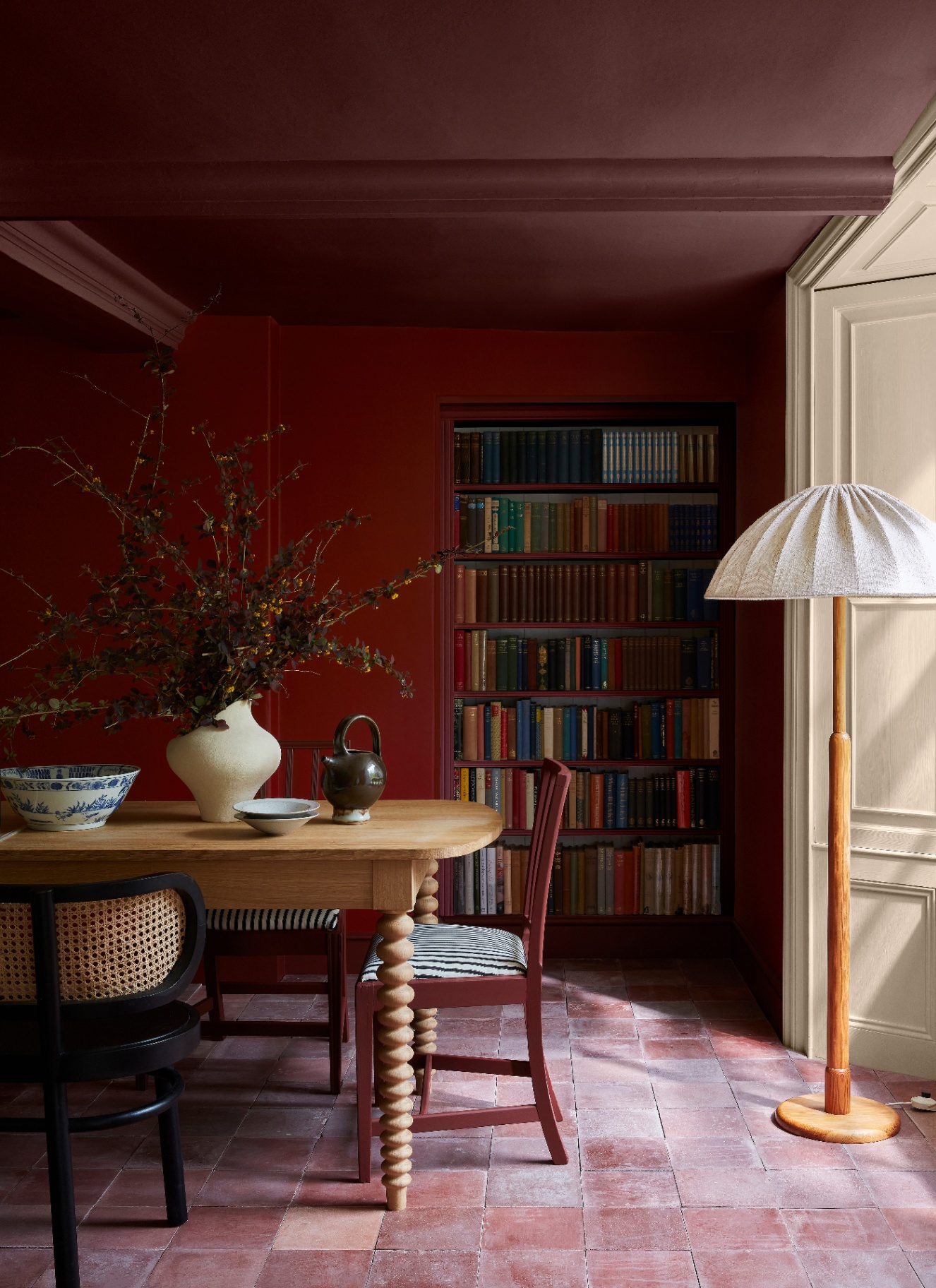 a dining room with a dark red colour drenched wall and ceiling, brick flooring, a bookshelf and a wooden bobbin floor lamp