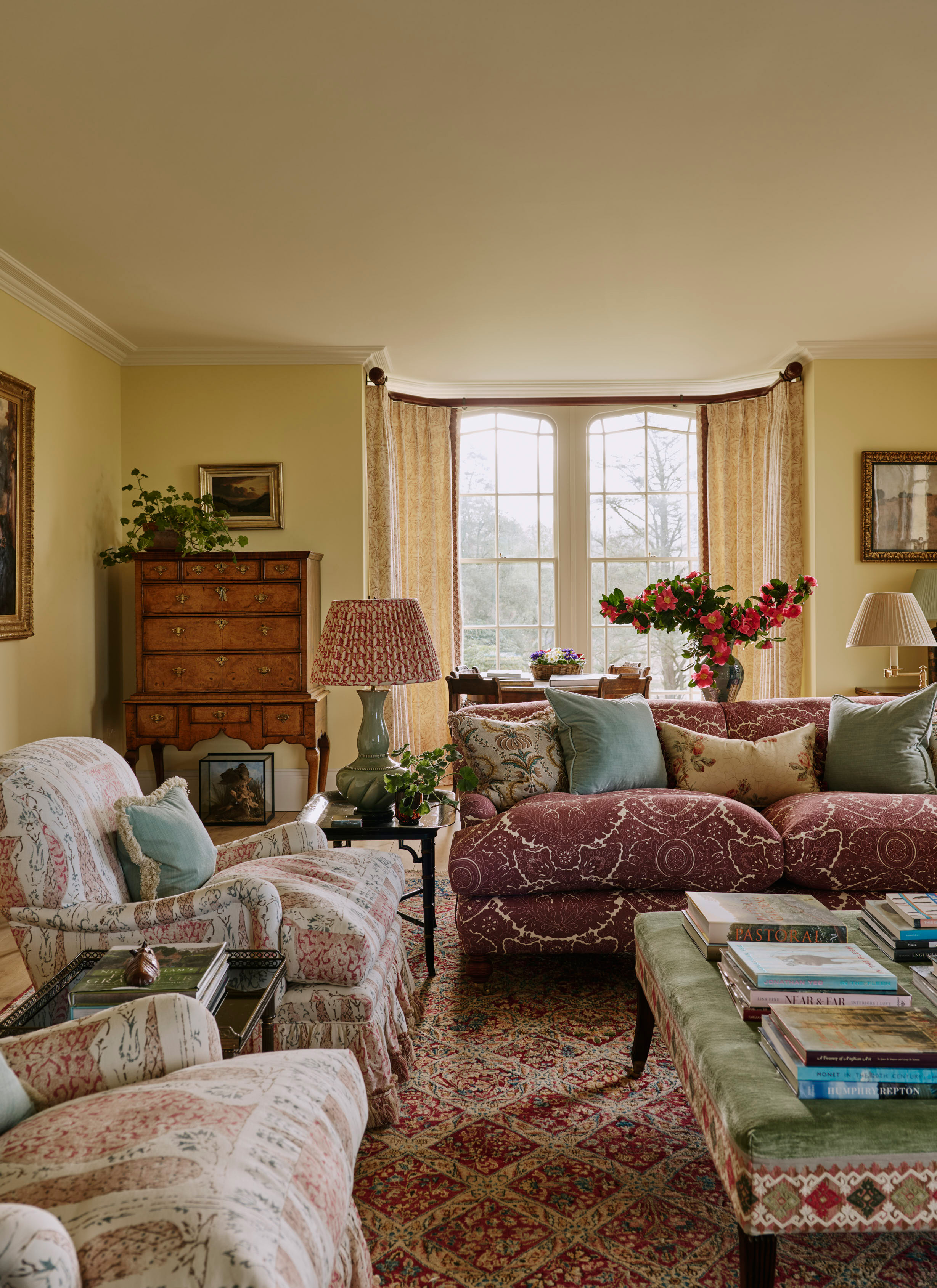An elegant English country living room featuring a patterned deep red sofa, floral upholstered armchairs, a green velvet ottoman stacked with art books, an antique tallboy cabinet, and large bay windows framed by patterned curtains.