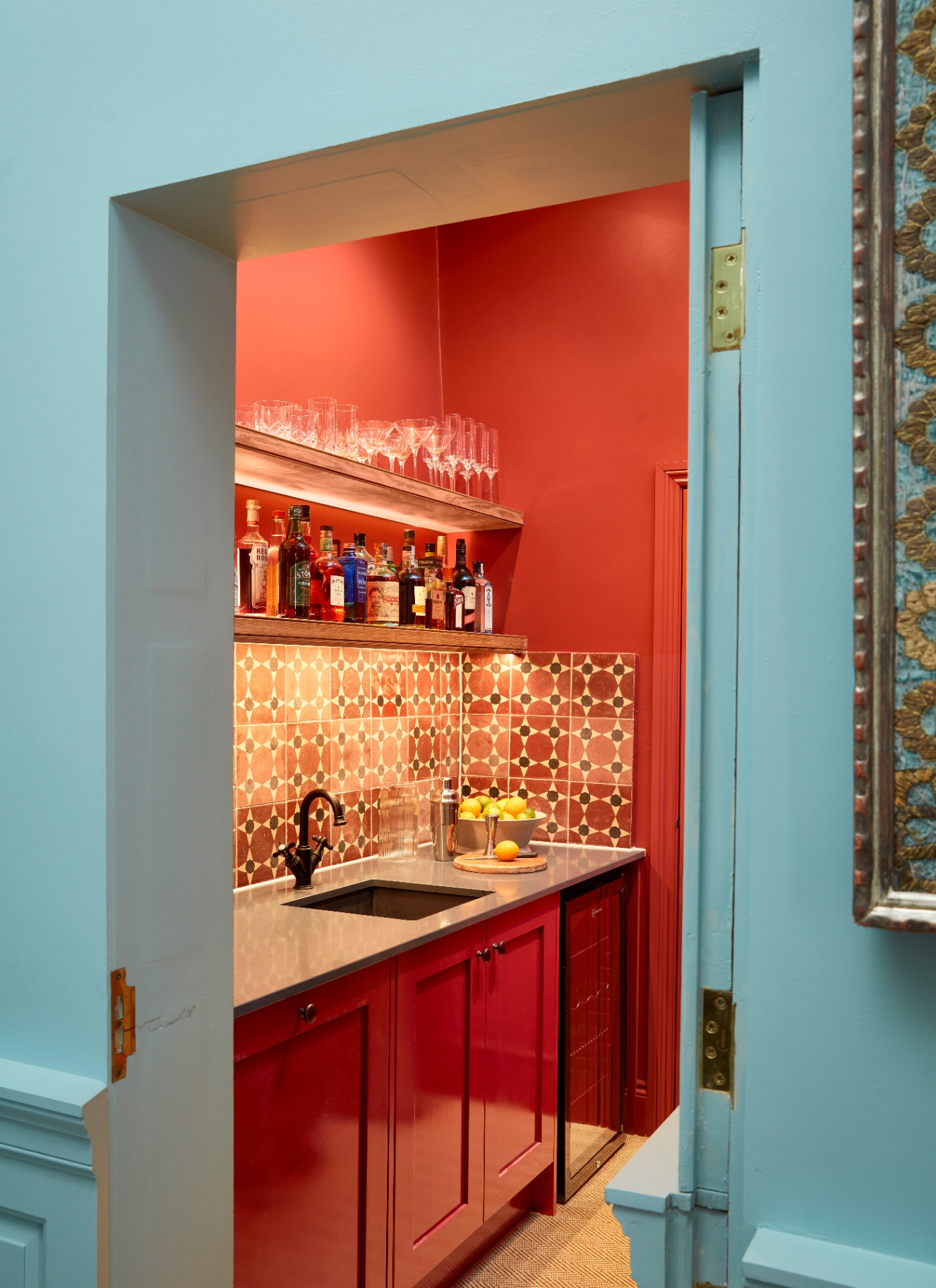 a bright turqoise wall with an open door looking into a small kitchen with bright red cabinets and shelving with glasses and bottles