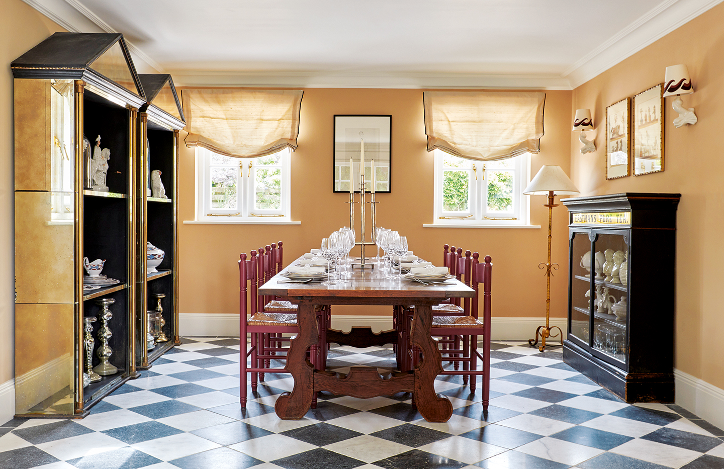 A dining room with checkerboard white and black marble floor. A large table sits in the centre with red gloss chairs. Two mirrored shelving units stand to the left and a slim display cabinet to the right. Relaxed blinds dress two windows on the back wall