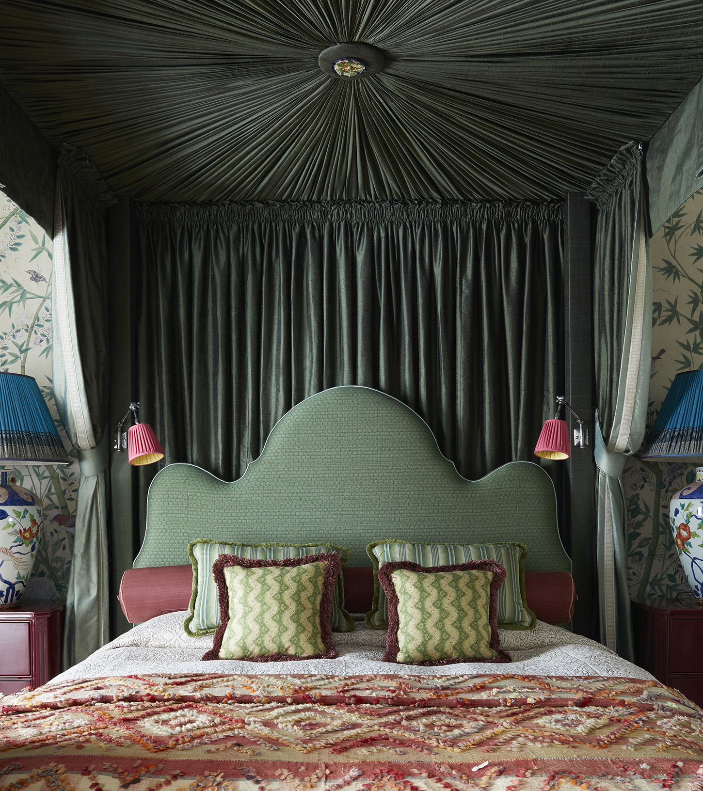 a four poster bed with a green pleated all over canopy, a pale green wavy headboard with two pink wall lights either side, green cushions, white bedding and a red patterned throw at the end of a double bed