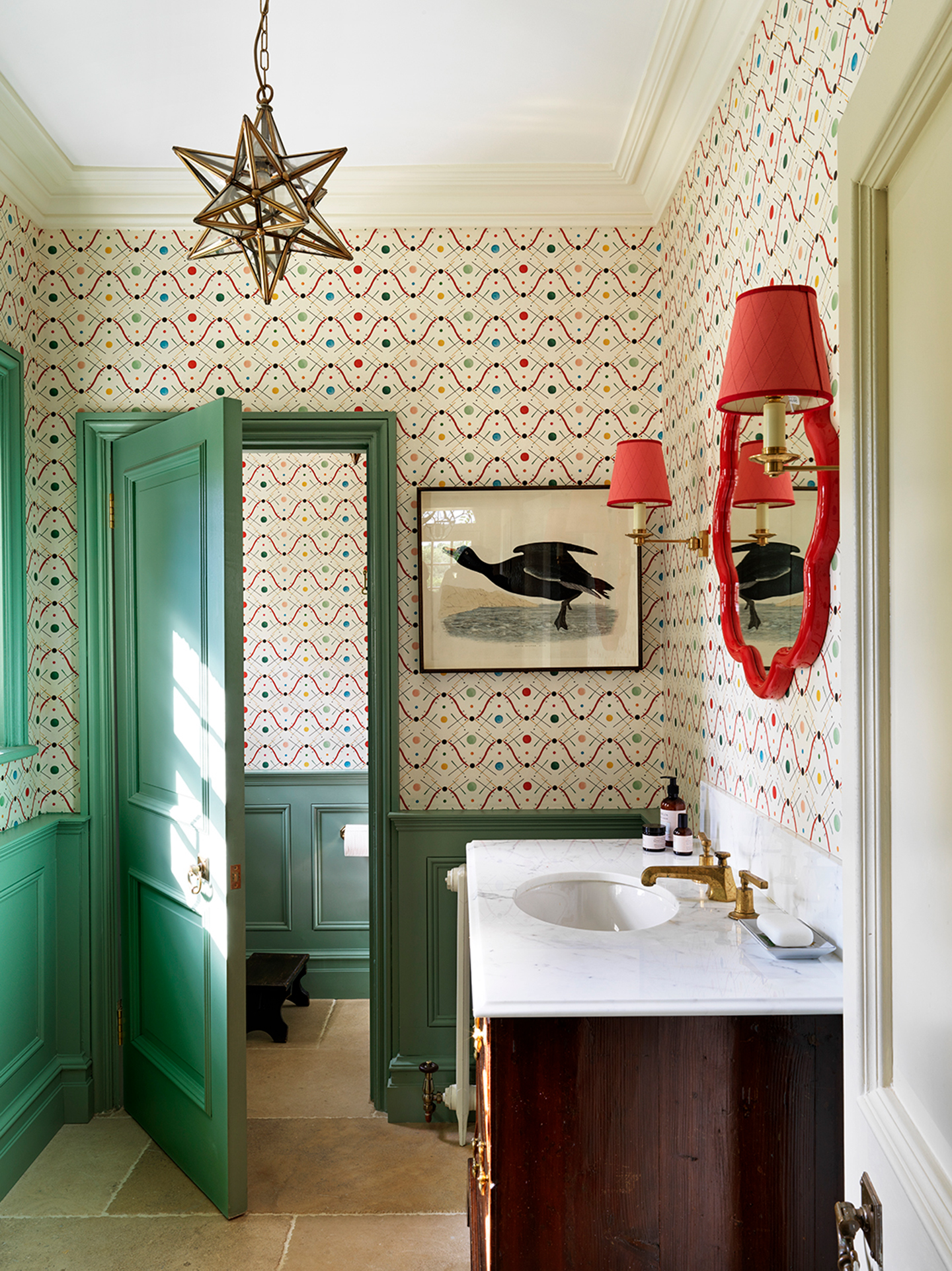 A small bathroom with graphic, colourful wallpaper on the upper half of the walls and painted green panelling on the lower half. A star shaped pendant hangs while wall lights with red shades and a red flower mirror hang above a wood-and-marble basin