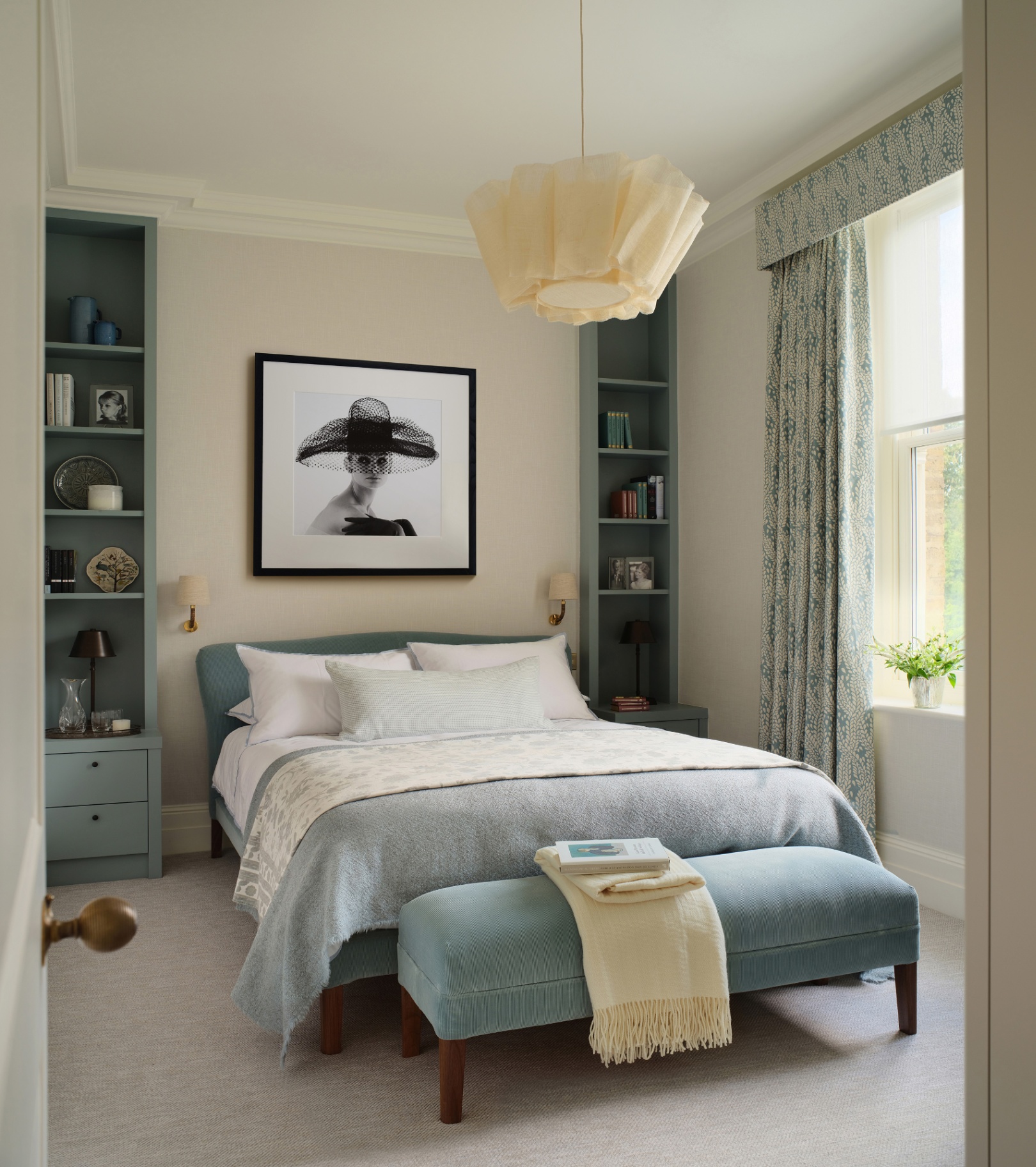 a bedroom with cream walls and blue painted built in book shelves either side of a double bed with white bedding and a blue bench at the foot of the bed