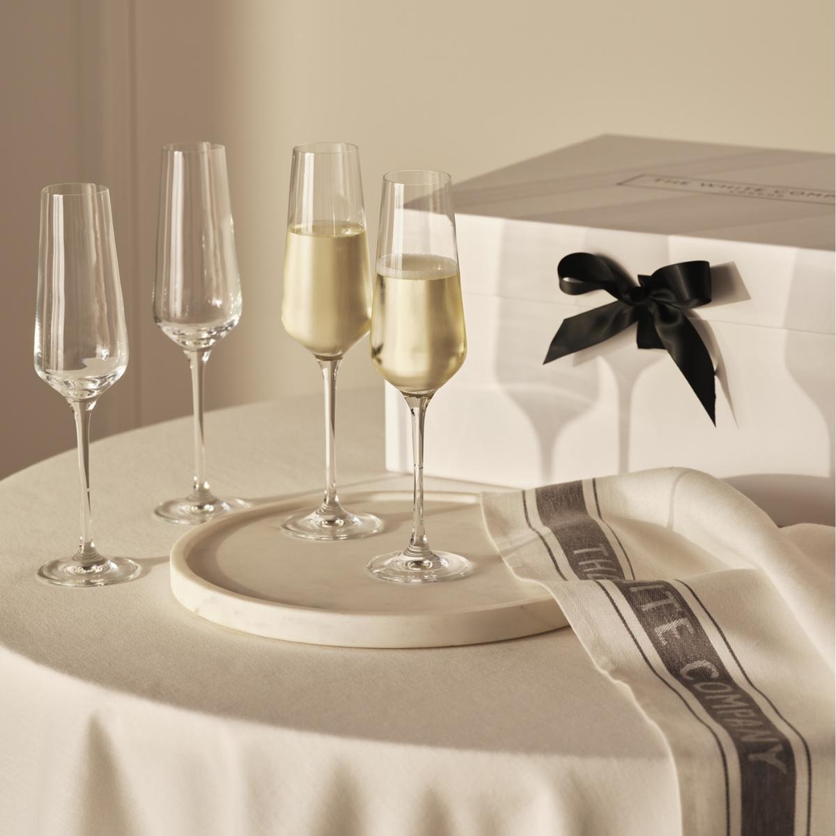 4 champagne glasses, a round marble tray, a white and navy tea towel and a white gift box with a black ribbon