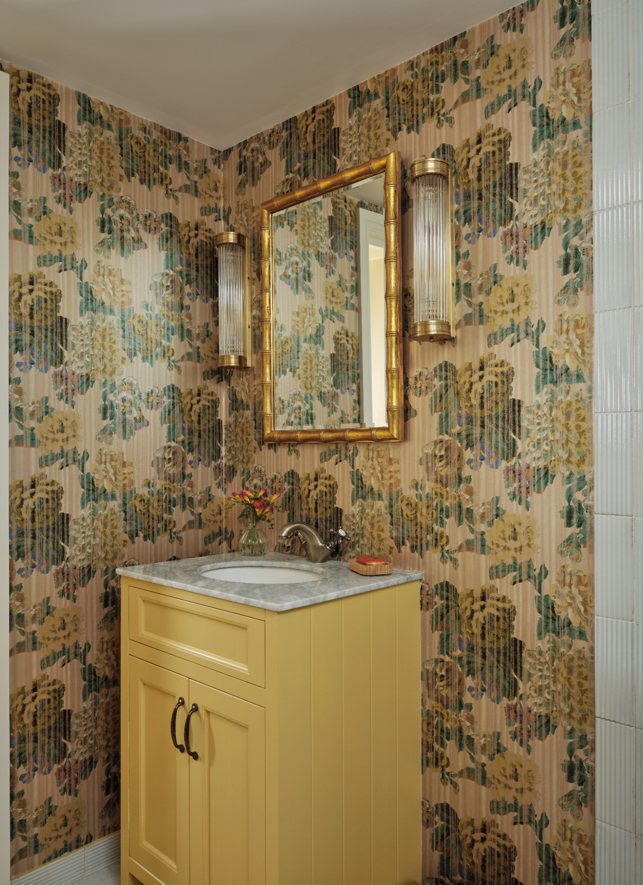a downstairs toilet with brown and yellow floral wallpaper, a yellow vanity with a marble top, a gold bamboo style mirror and two fluted glass sconces