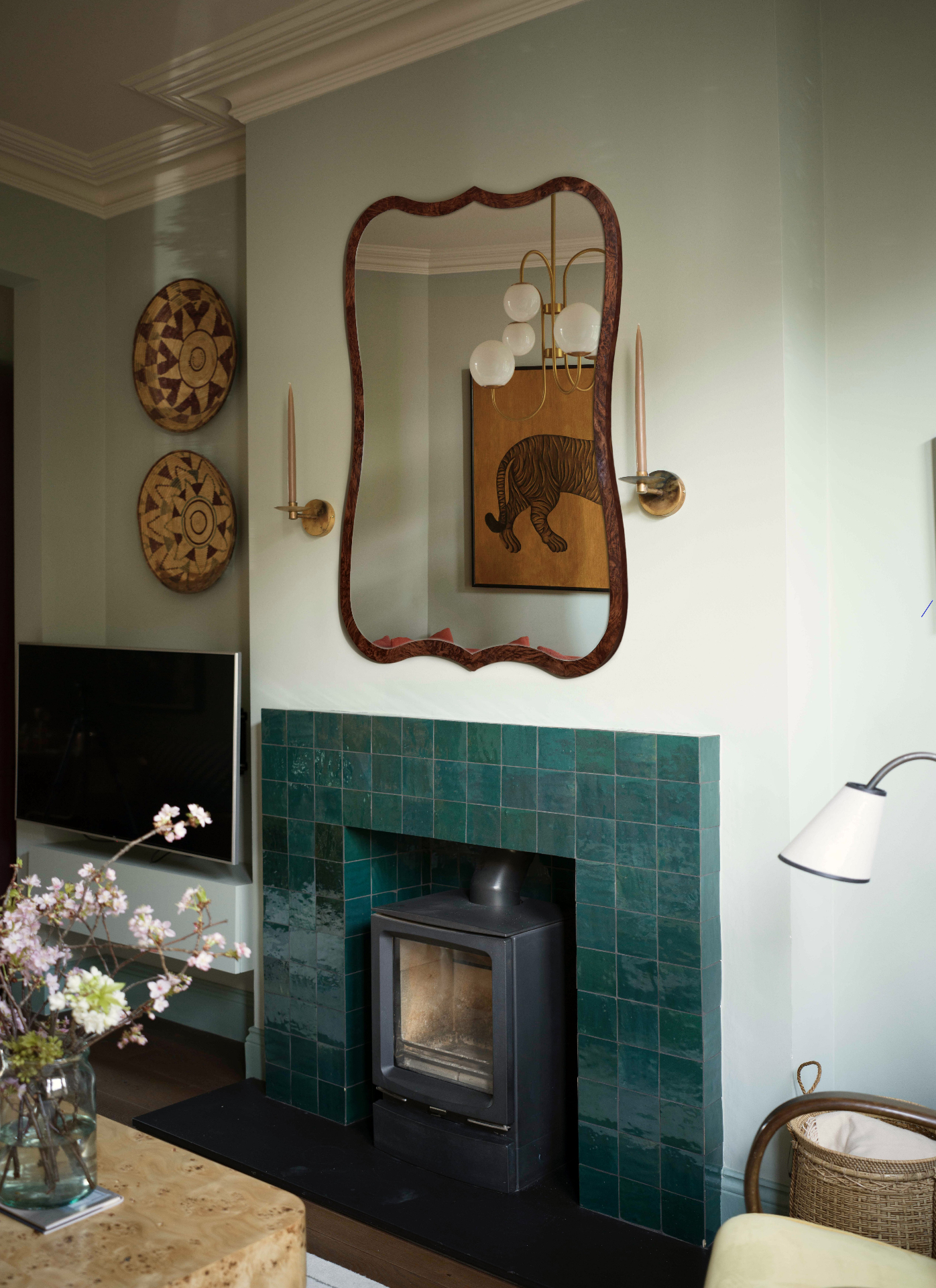 A living room corner with mint green walls, a teal green tiled fireplace, a large wavy edge wooden mirror above the fireplace reflecting a paitning of a tiger and two brass candle sconces either side