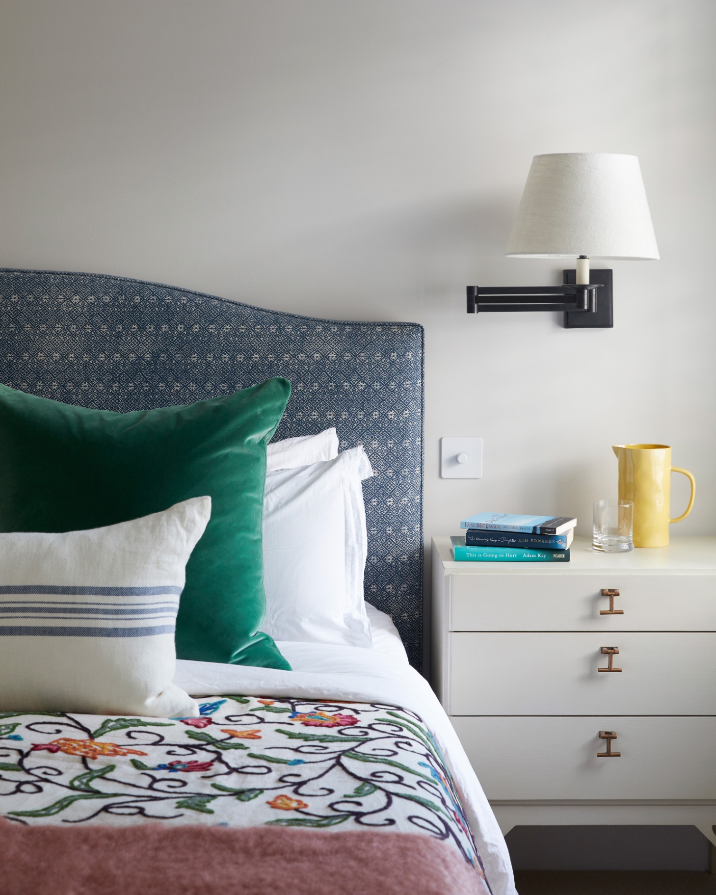 a close up of a bedroom corner with white walls, a white bedside table with a yellow water jug and a bed with a blue patterned headboard and a green velvet cushion