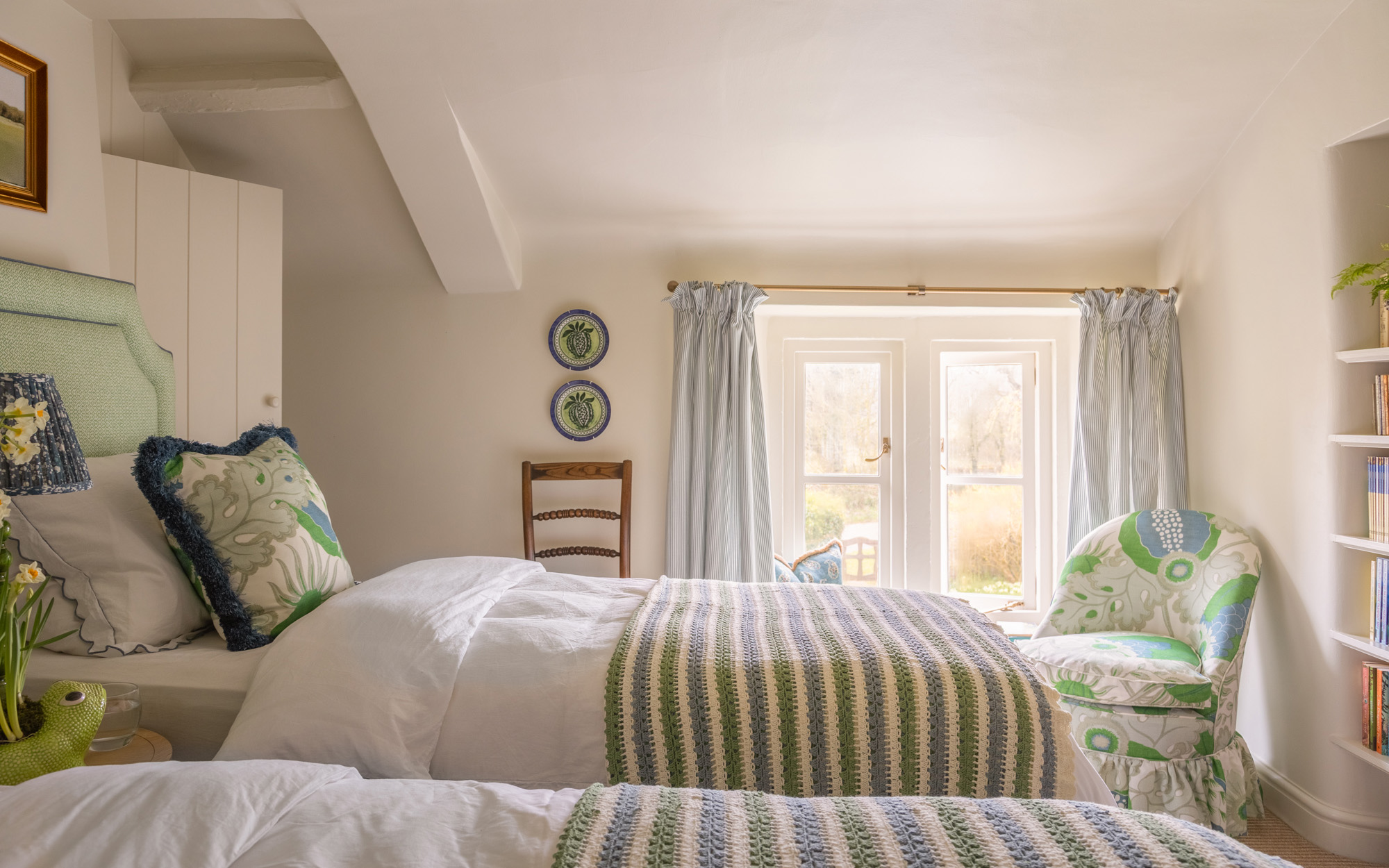 a cottage bedroom with white walls and wooden beams, a blue, green and white floral fabric skirted armchair in front of the window and two twin beds with white bedding and a navy and green and white bedspread on the end