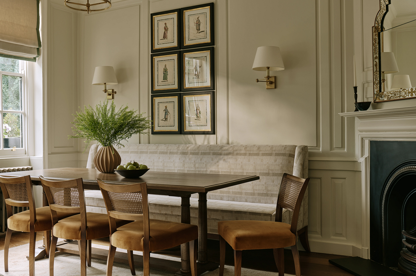 A wooden dining table is surrounded by four upholstered and cane dining chairs and a long upholstered bench, and is topped with a bowl of fruit and greenery in a vase. The wall behind is panelled and decorated with a pair of wall lights and artworks 
