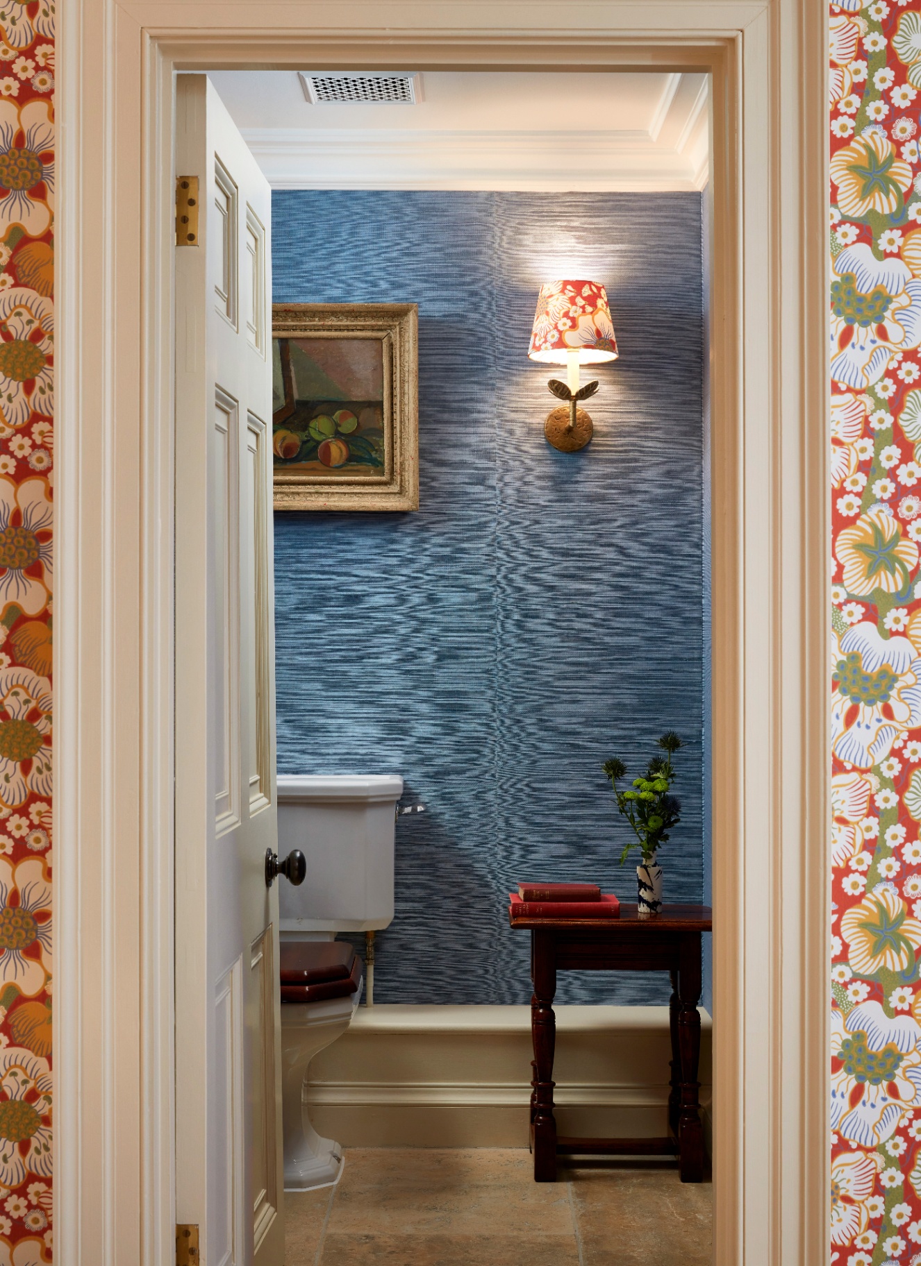 a downstairs toilet with blue textured wallpaper, a sconce with a red patterned shade, a white toilet with a lanscape paitning in a gold frame above it and a small dark wooden table with books and a plant