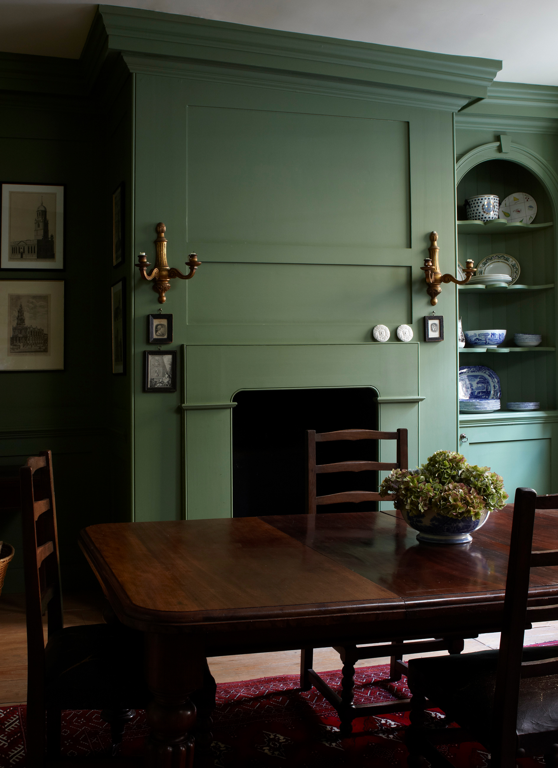 Moody olive green painted dining room with full wall panelling, built-in display arch, dark wooden dining table, brass sconces, and architectural prints