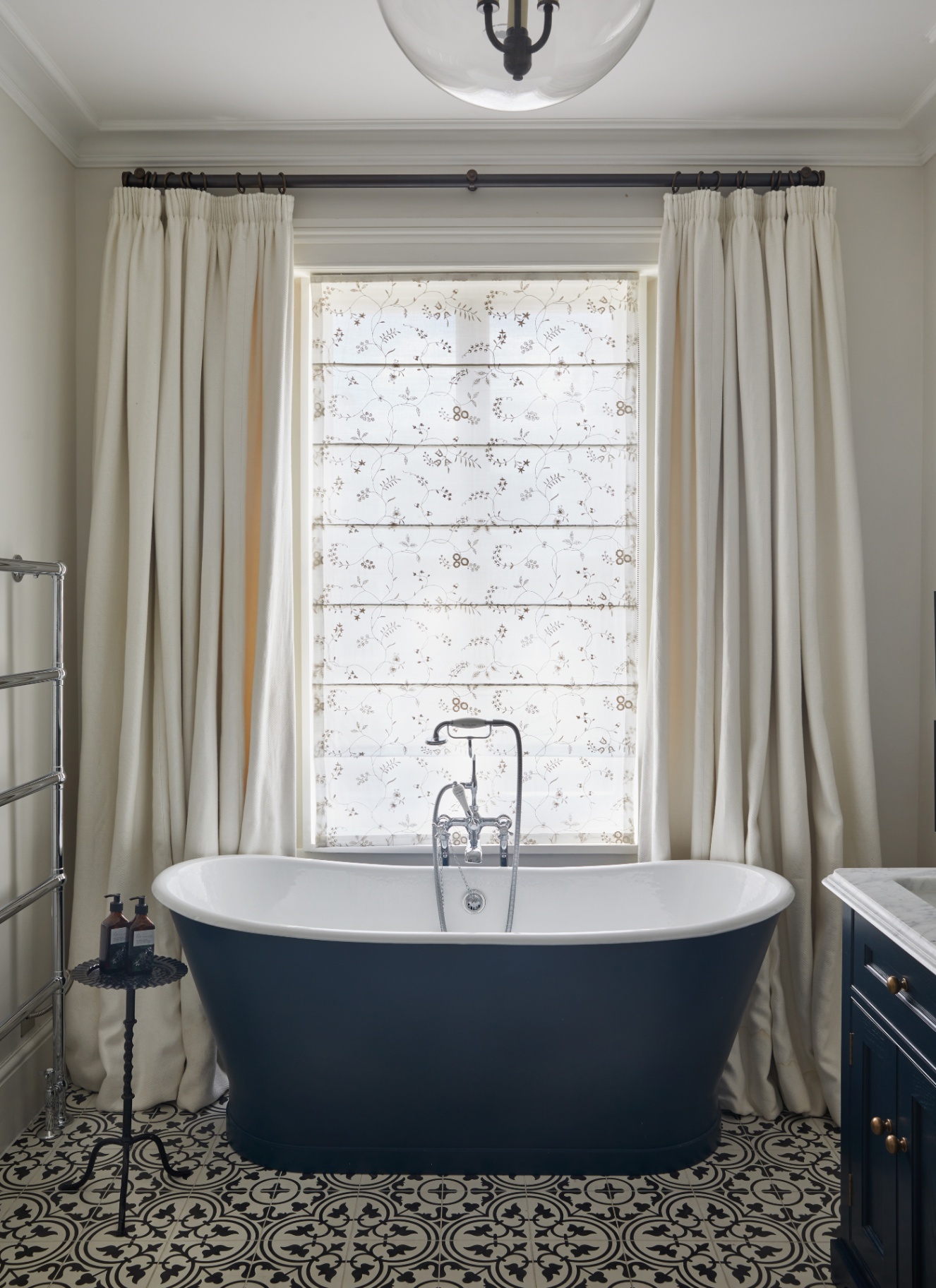 a bathroom with a dark blue freestanding bath tub, black and white patterned floor tiles and a window with long cream curtains
