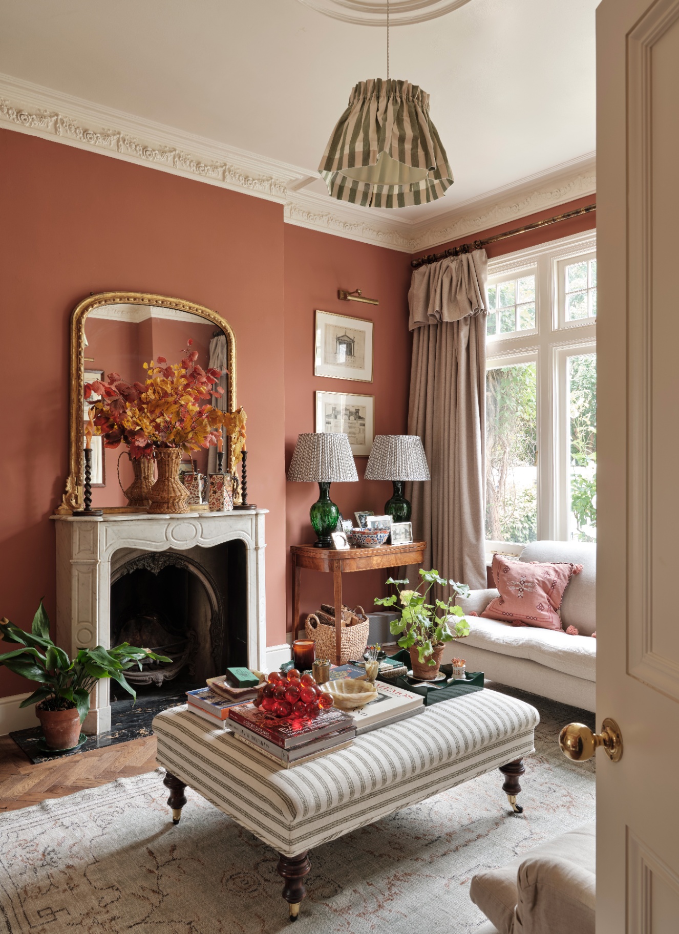 a living room with teracotta walls, a marble fireplac with a vase of autumm leaves above it and a white and beige striped ottoman