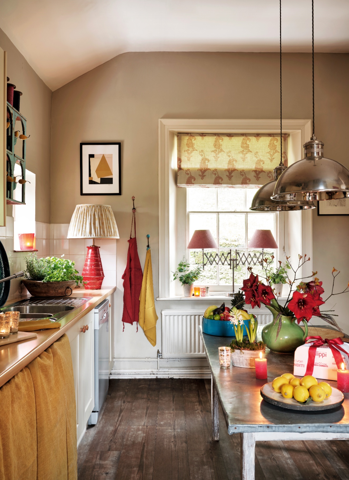 A kitchen island with a green pitcher of crimson amaryllis, potted hyacinths, fruit, and candles, with a sink, a lamp, and hanging aprons in the background.