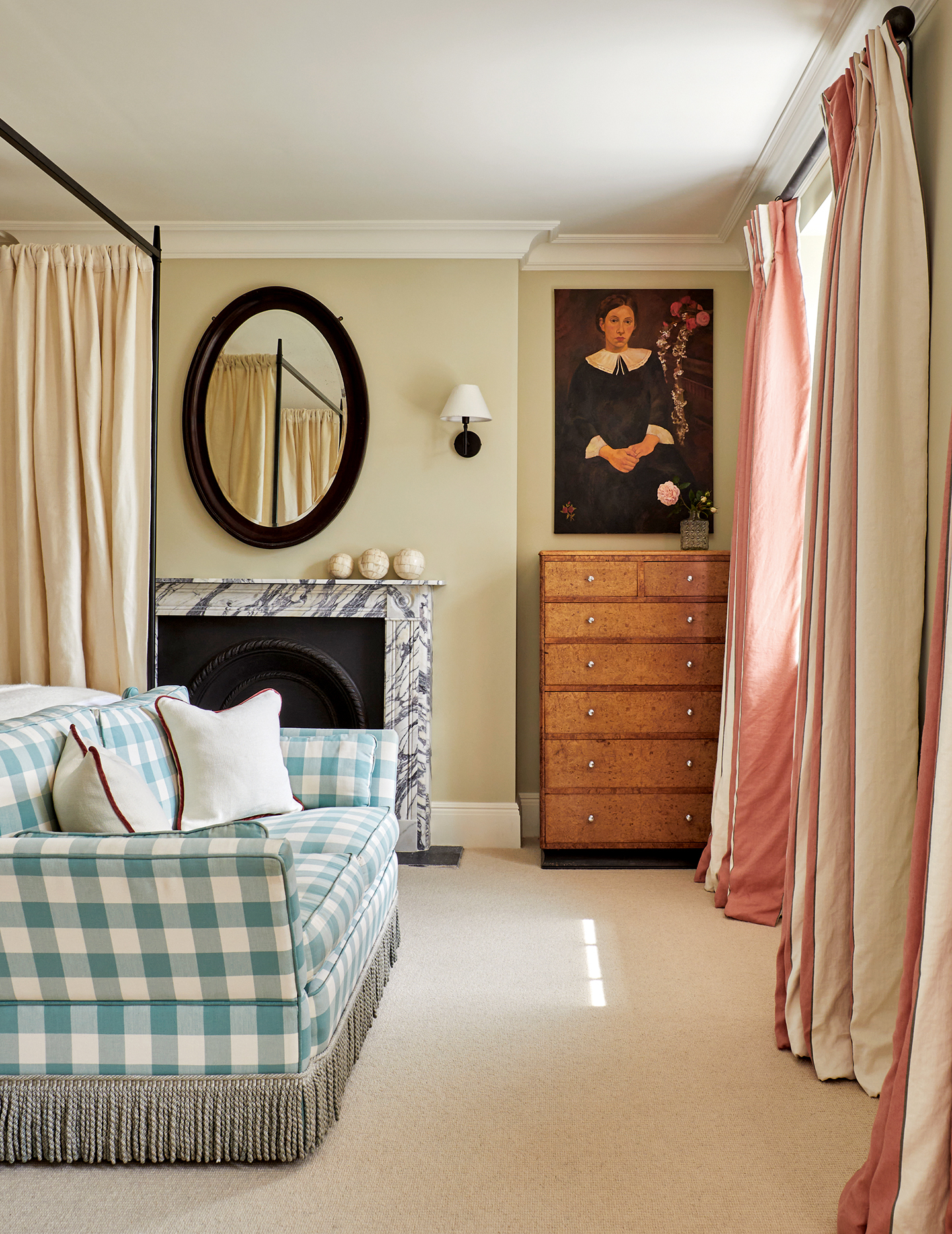 A fringed, blue and white check sofa sits at the end of a metal four-poster bed in a light green bedroom. A marble fireplace and tall burl wood chest of drawers sit against the back wall with an oval mirror and large portait painting above