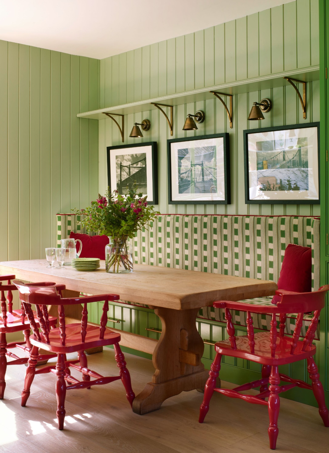 Sage green panelled dining nook featuring a bench with green patterned cushions, red wooden chairs, and brass wall sconces above artwork.