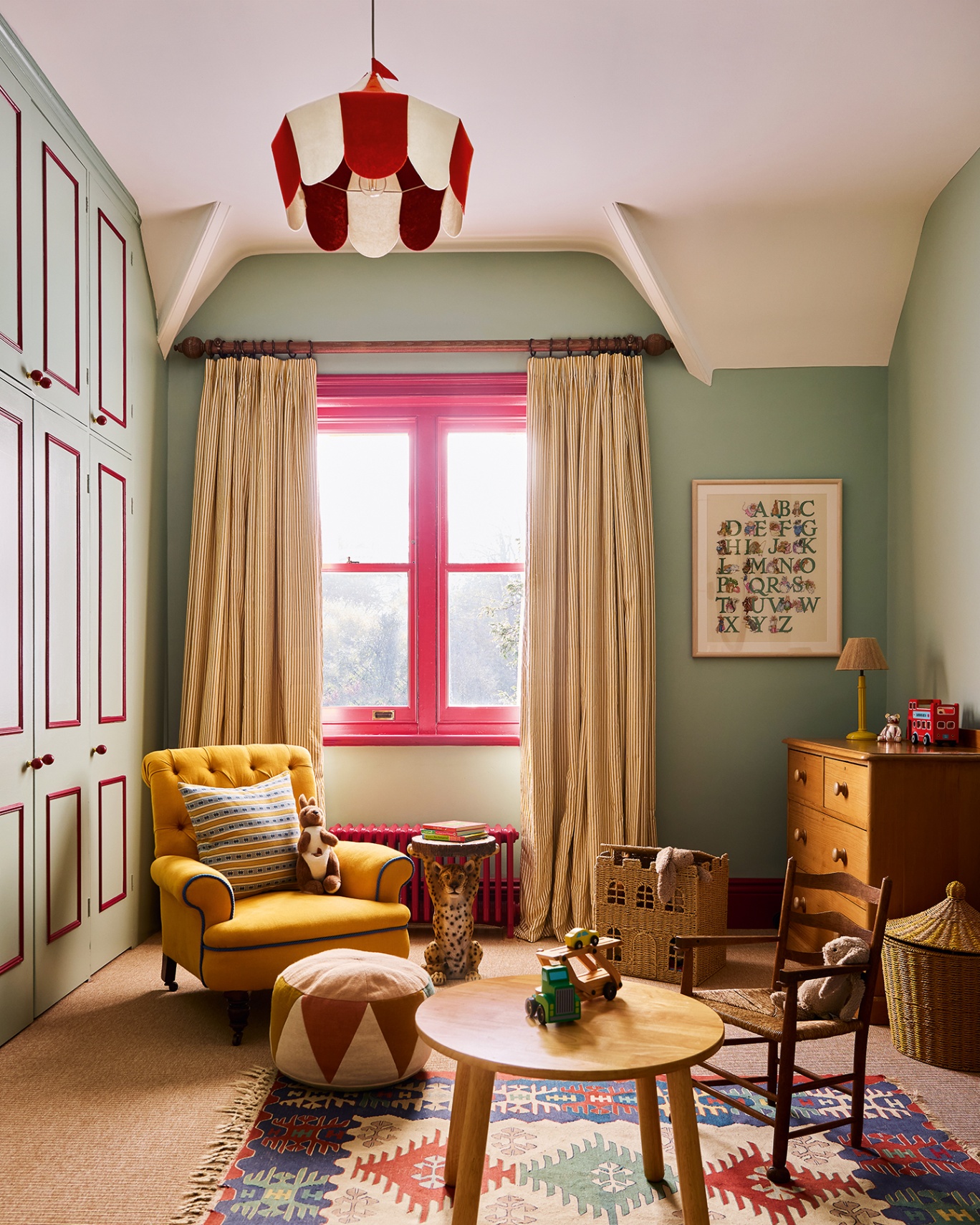A playroom with bllue walls and red woodwork and radiator. In the foreground is a patterned rug, mini wooden circular table, mini wooden chair, colourful pouffe and yellow armchair. To the right is a wooden chest of drawers and alphabet artwork on the wall