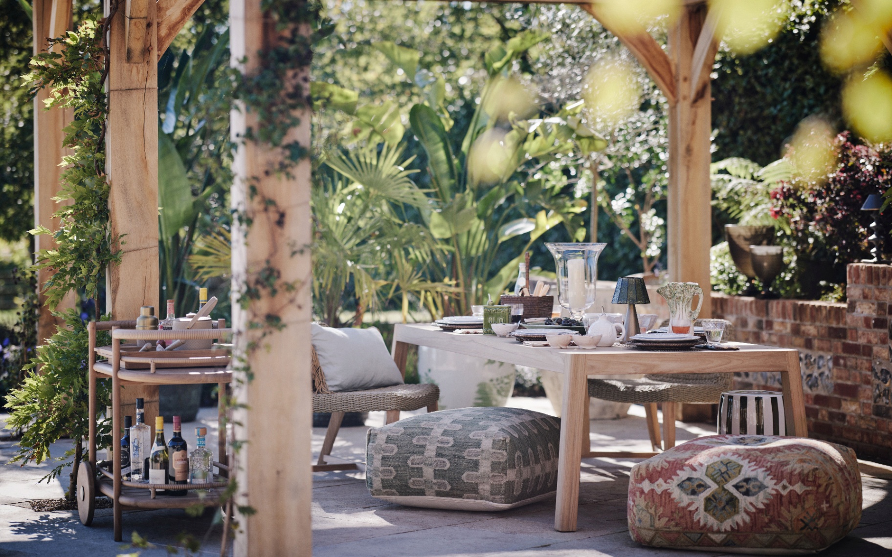 an outdoor dining space under a wooden pergola on a grey patio with a wooden bar cart next to it and lots of green plants behind the table