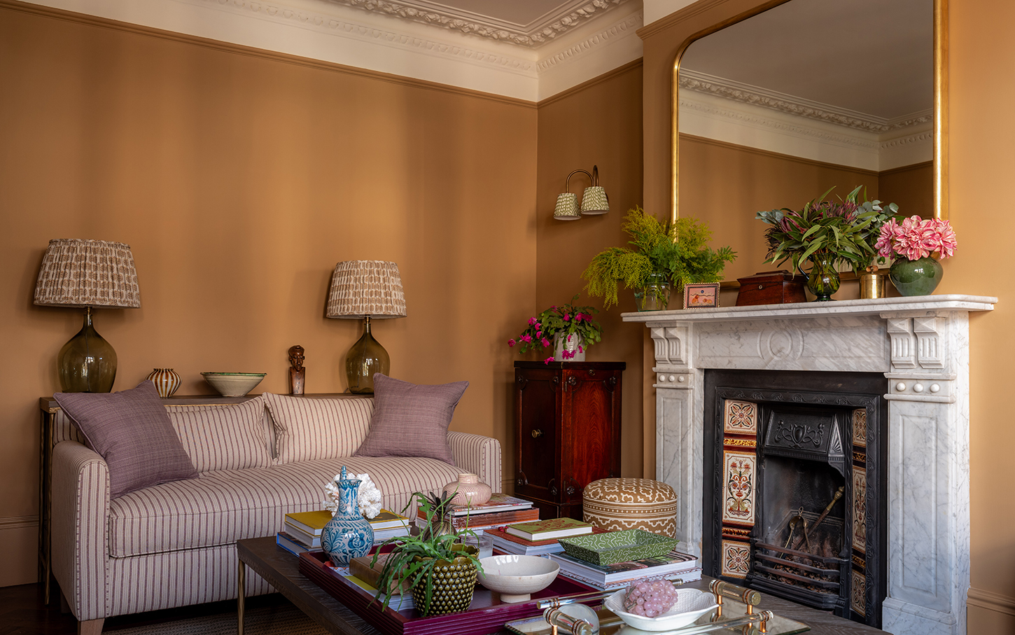 A striped purple sofa with purple linen cushions in a living room with caramel brown walls, behind it is a pair of statement table lamps with patterned fabric shades and to the right is a Victorian marble fireplace with a large antique gold mirror above