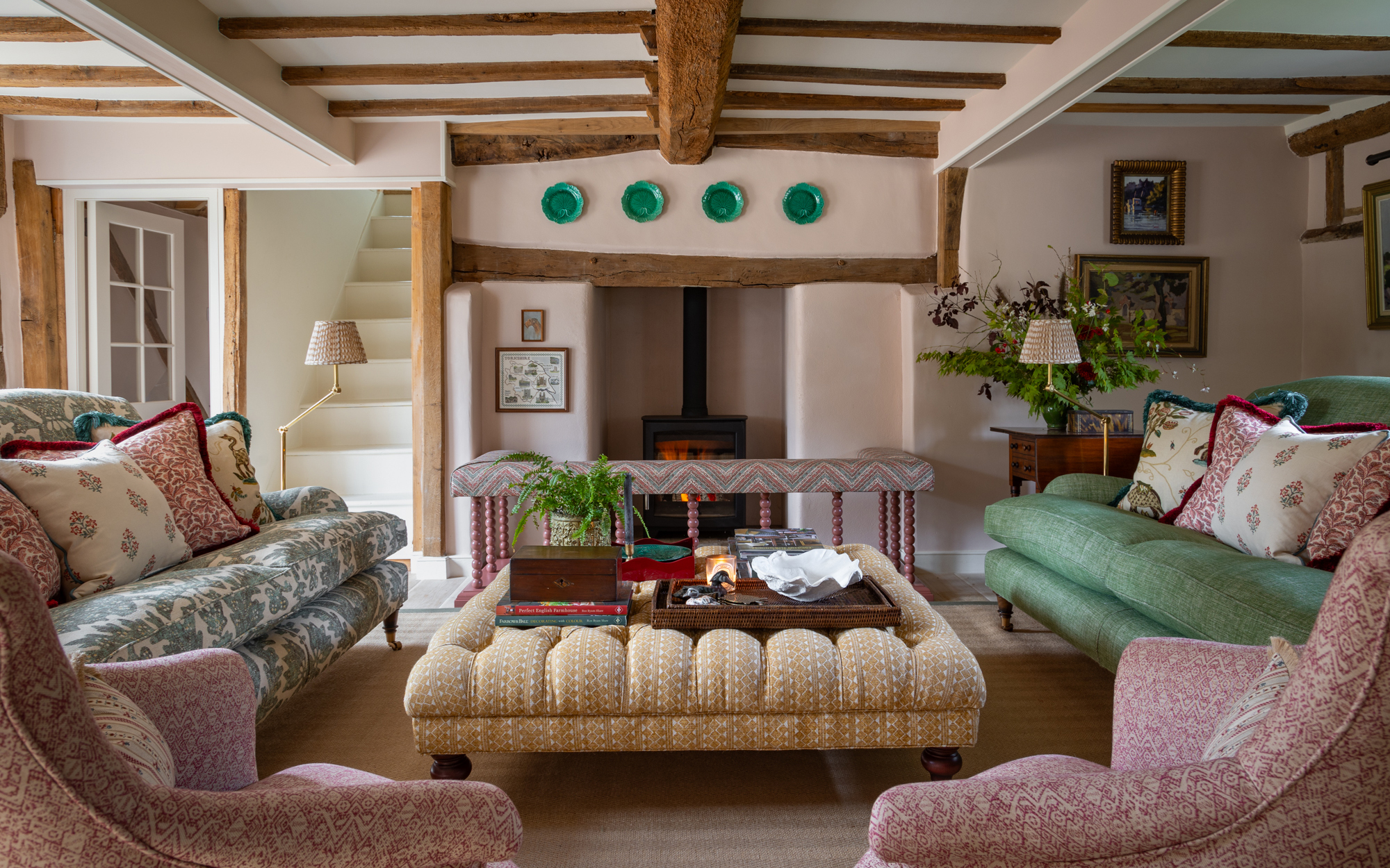 a living room with wooden beams, a black wood burner with green plates hanging on the wall above it and sofas and armchairs of different colour facing a yellow ottoman in the centre