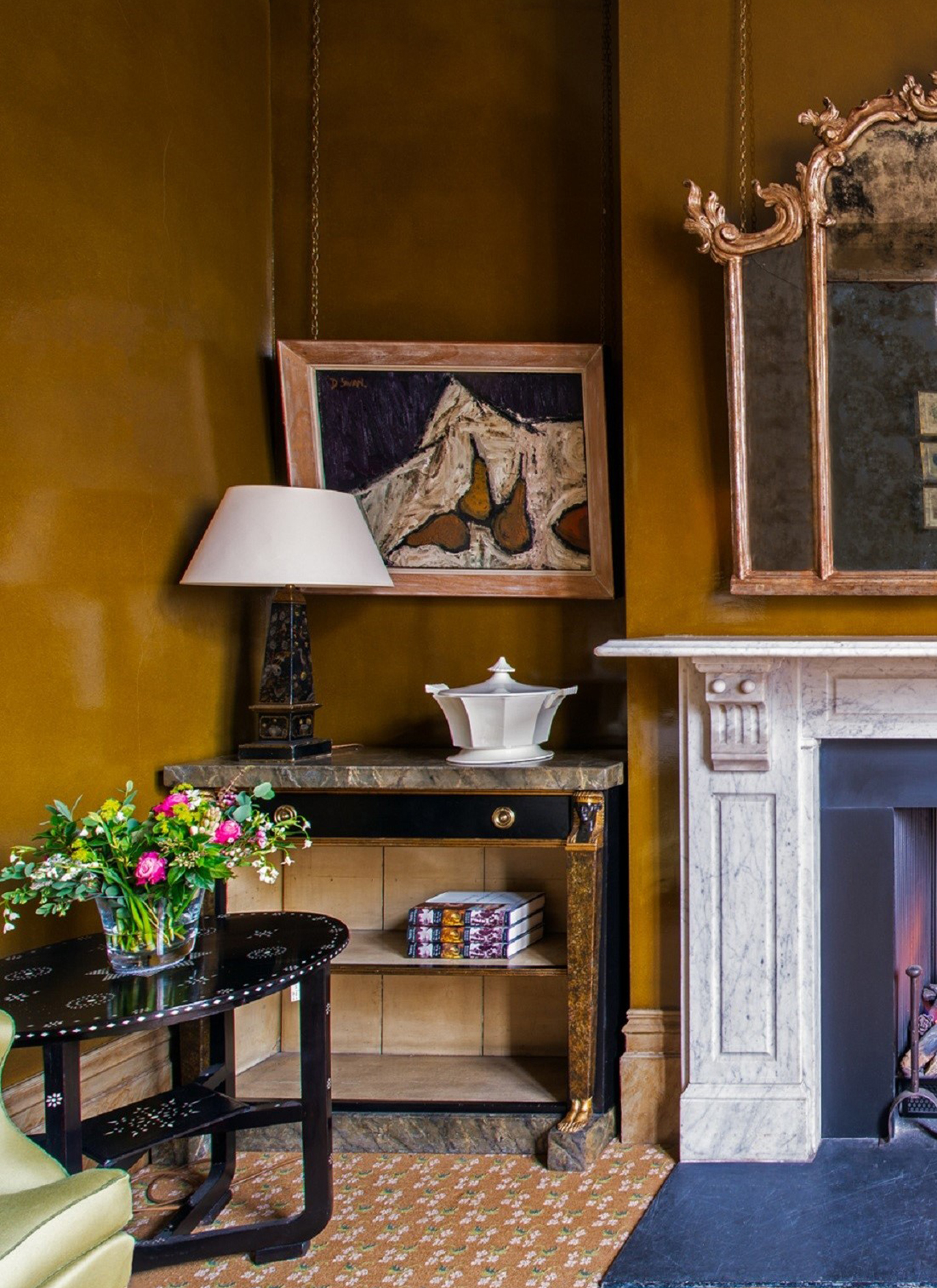 a living room corner with dark ochre walls, a large white marble fireplace, a marble side table with an abstract horse painting above, orange patterned carpet and a small dark round side table with pink flowers