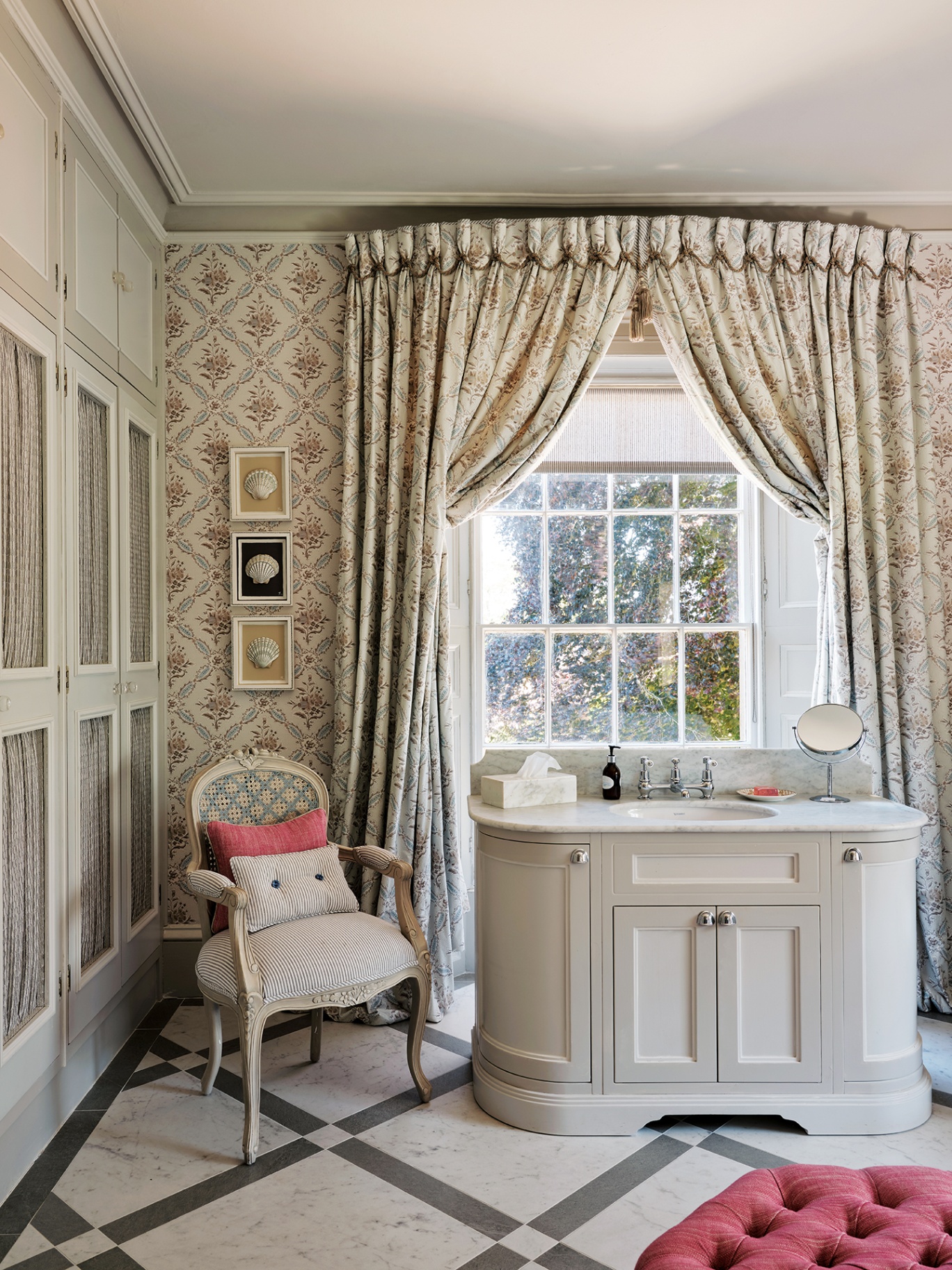 a close up of a bathroom with floral wallpaper, floral curtains and a diamond-patterned stone floor. A curved vanity unit with a marble top sits in front of the window with a small antique chair and built in wardrobes next to it