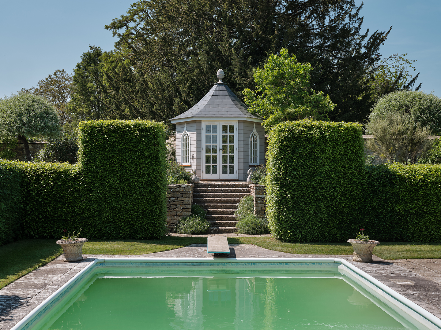 a hexagonal summer house with double doors and arched windows sits atop stone steps in between large sculpted hedges, behind a large swimming pool