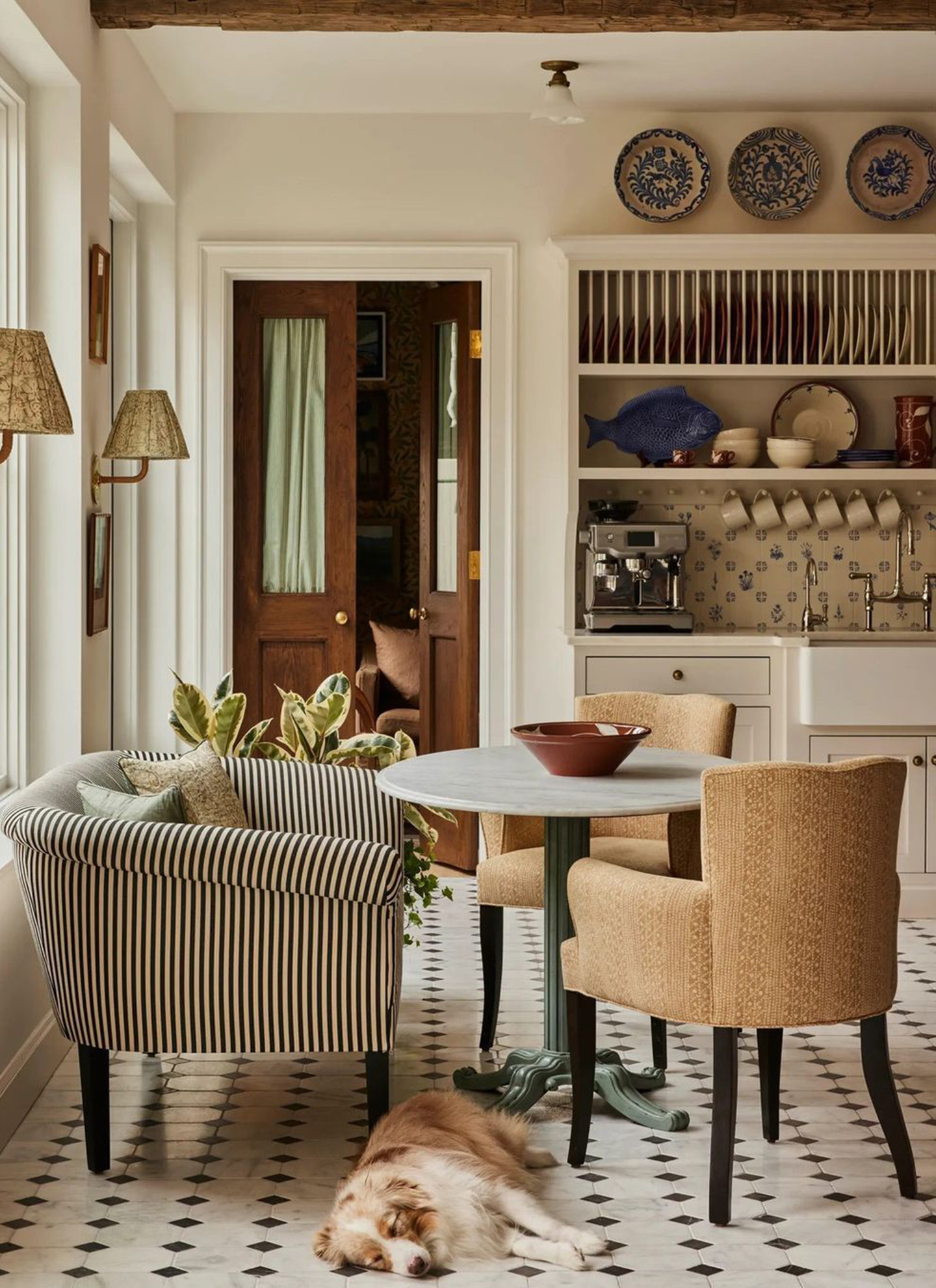 Parisian bistro-style dining nook with a marble table, striped armchair, sleeping dog on tiled floor, and a countertop espresso machine setup beside a butler sink