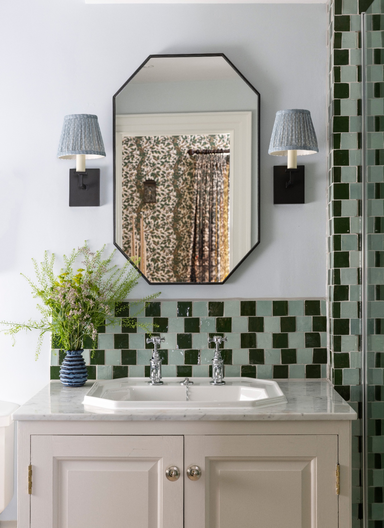 a bathroom sink area with an octanganol mirror, two pleated sconces, green checkerboard tiles and a cream and marble vanity unit