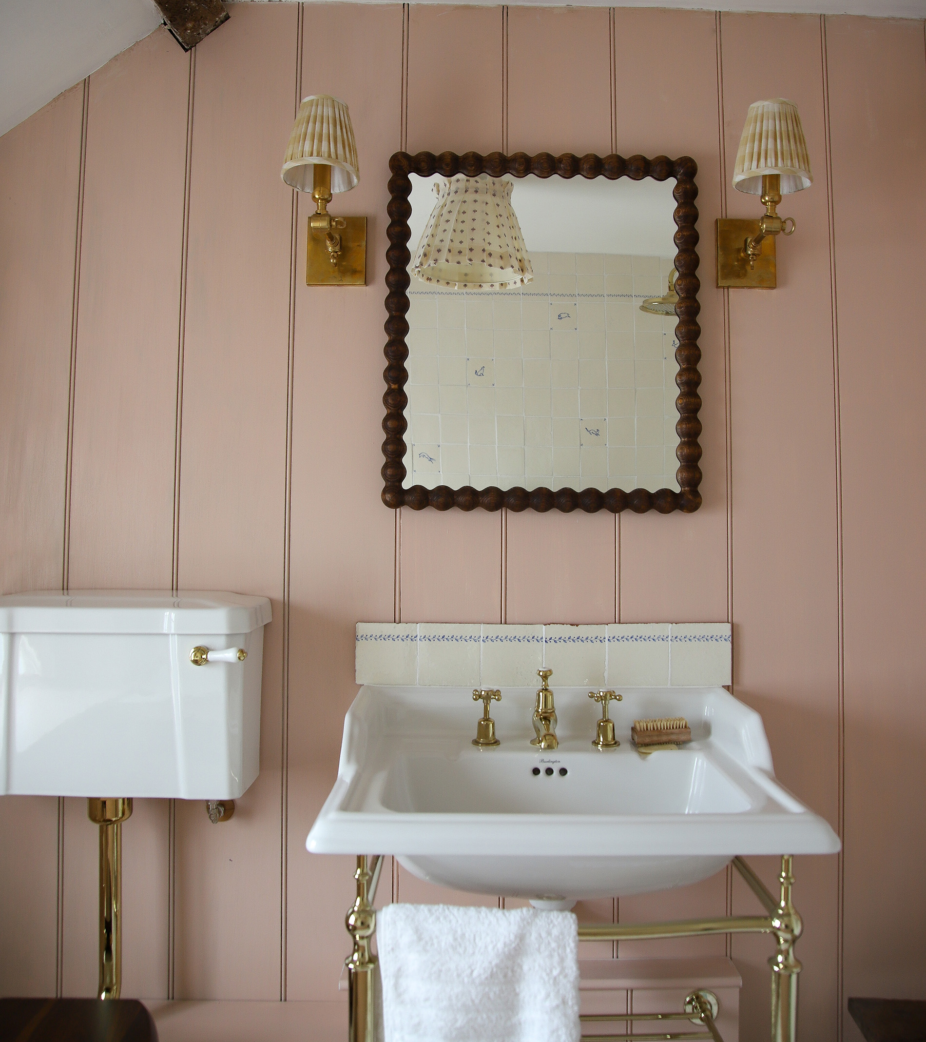 marlborough tiles pale pink bathroom with a gold and white victorian basin, a dark brown bobbin rectangluar mirror with two yellow and white pleated sconces either side