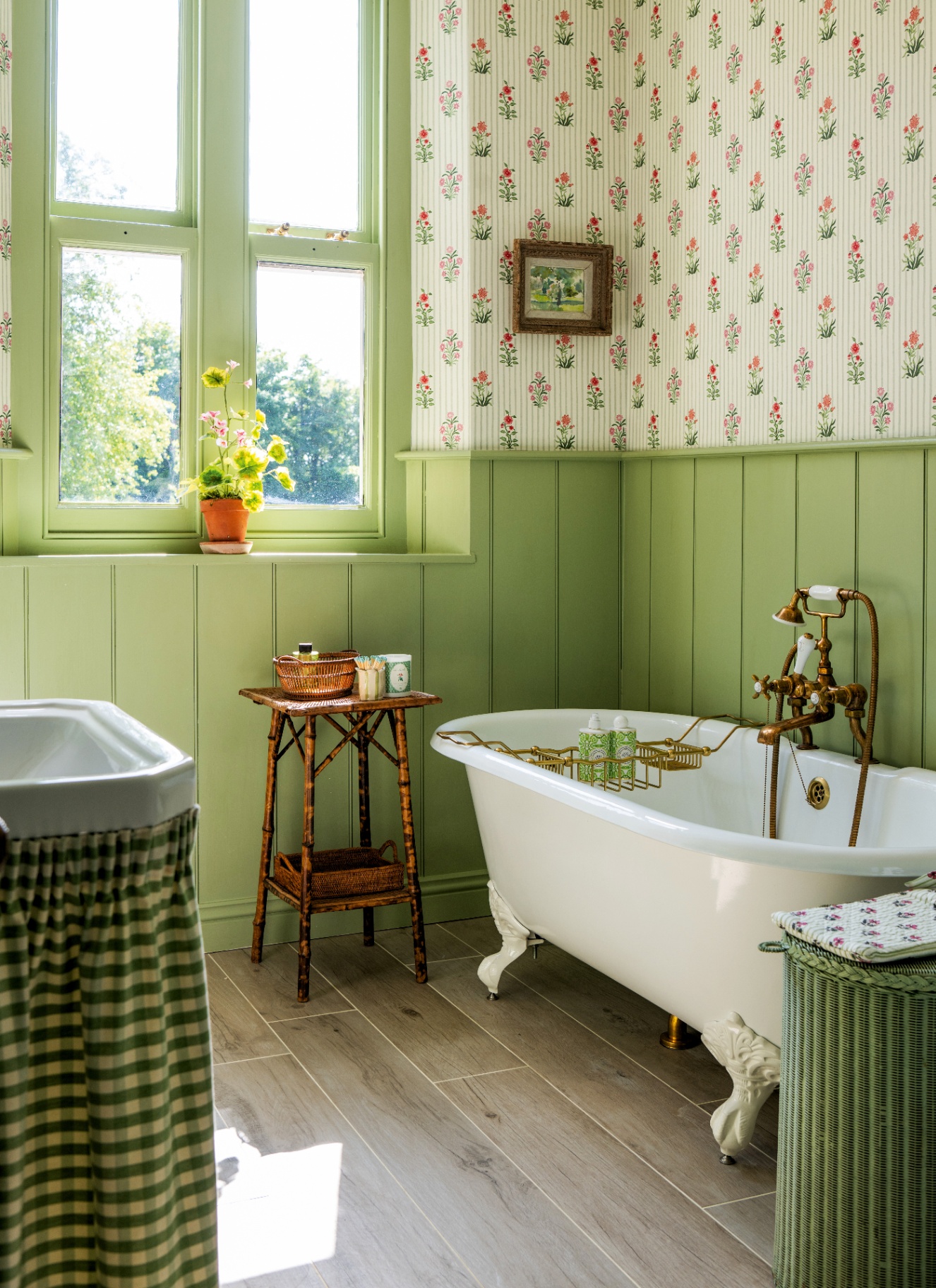 Meadow green bathroom with a white clawfoot bathtub, brass fixtures, and green vertical beadboard panelling below floral wallpaper. A bamboo side table sits near a bright window, alongside wood-look tile flooring and a green gingham skirted vanity