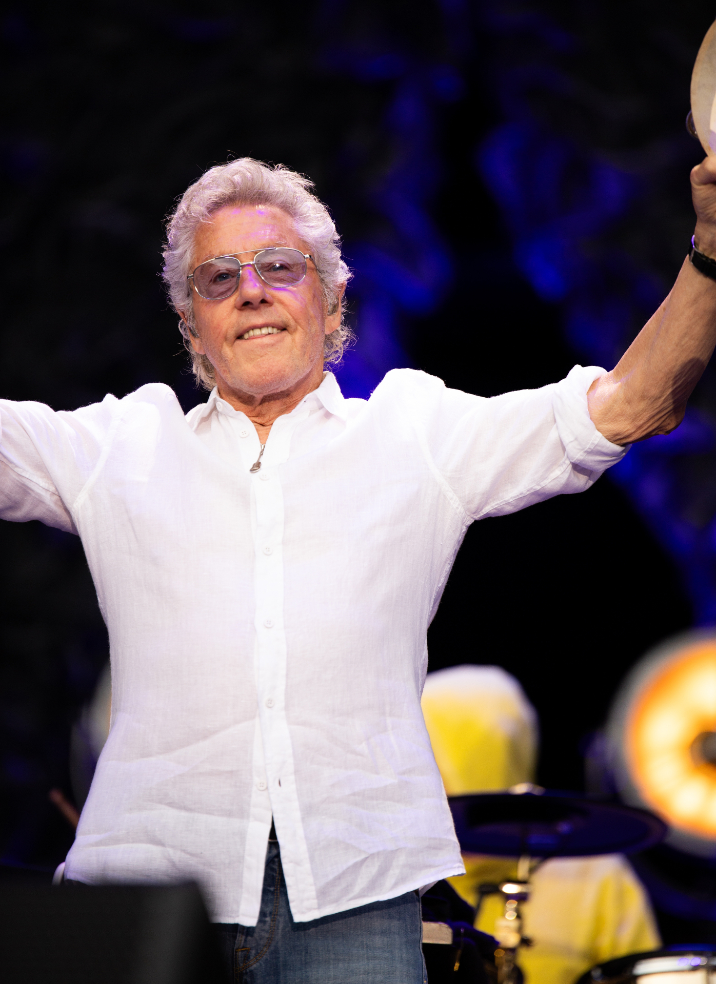 Roger Daltrey wearing a white shirt and glasses holding a tambourine on stage