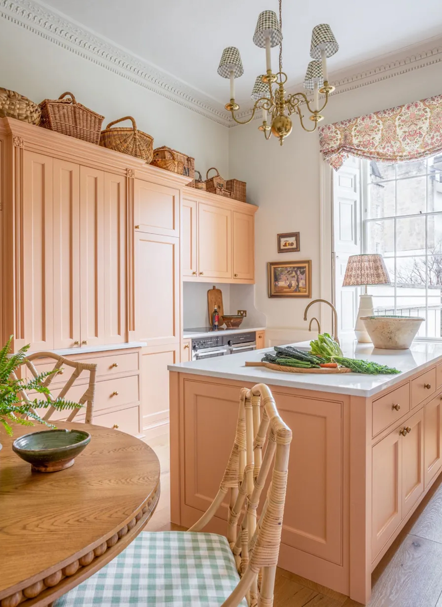a kitchen with salmon pink cabinets, a wooden round table with cane chairs, a gold chandelier with gingham shades and a collection of wicker baskets on top of the cabinets