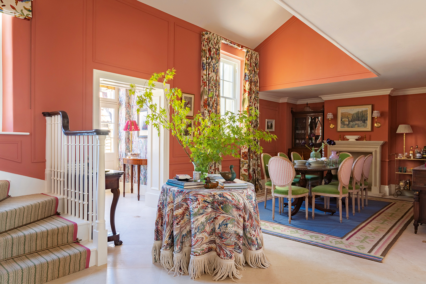 A bold orange dining space with an antique table and green upholstered chairs sat on a colourful, floral rug in front of a fireplace. A circular table with a marbled tablecloth and stairs with a striped runner can be seen in the foreground