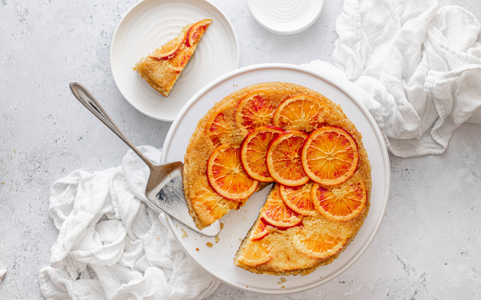 Overhead view of a round orange upside-down cake topped with translucent, caramelized blood orange slices arranged on a white ceramic cake stand, with a stainless steel cake server lifting a slice beside a small plate holding a single piece.