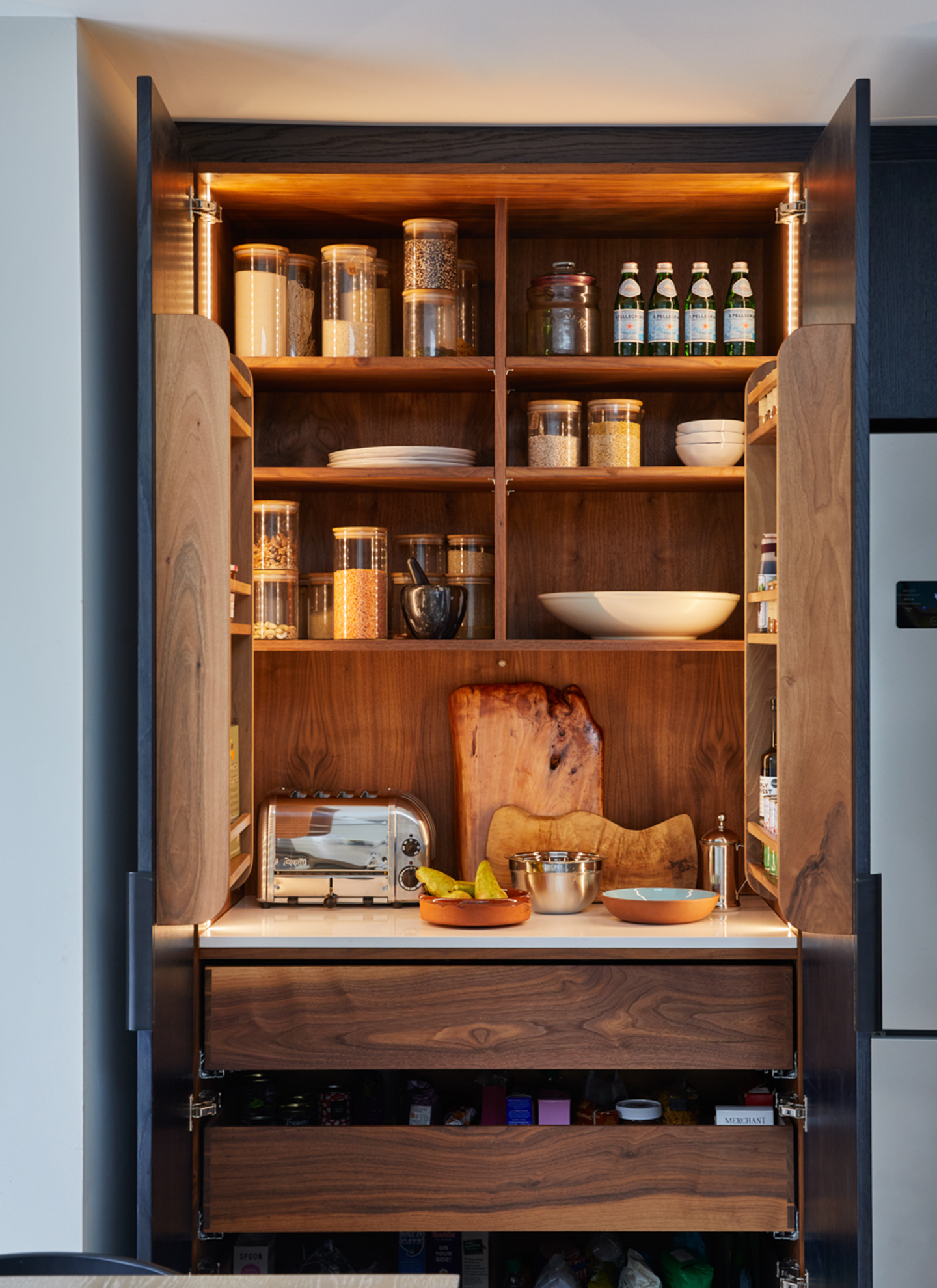 Open walnut larder cupboard with warm LED backlighting, integrated shelving for glass storage jars, a marble countertop with a chrome toaster, and wooden drawers below.